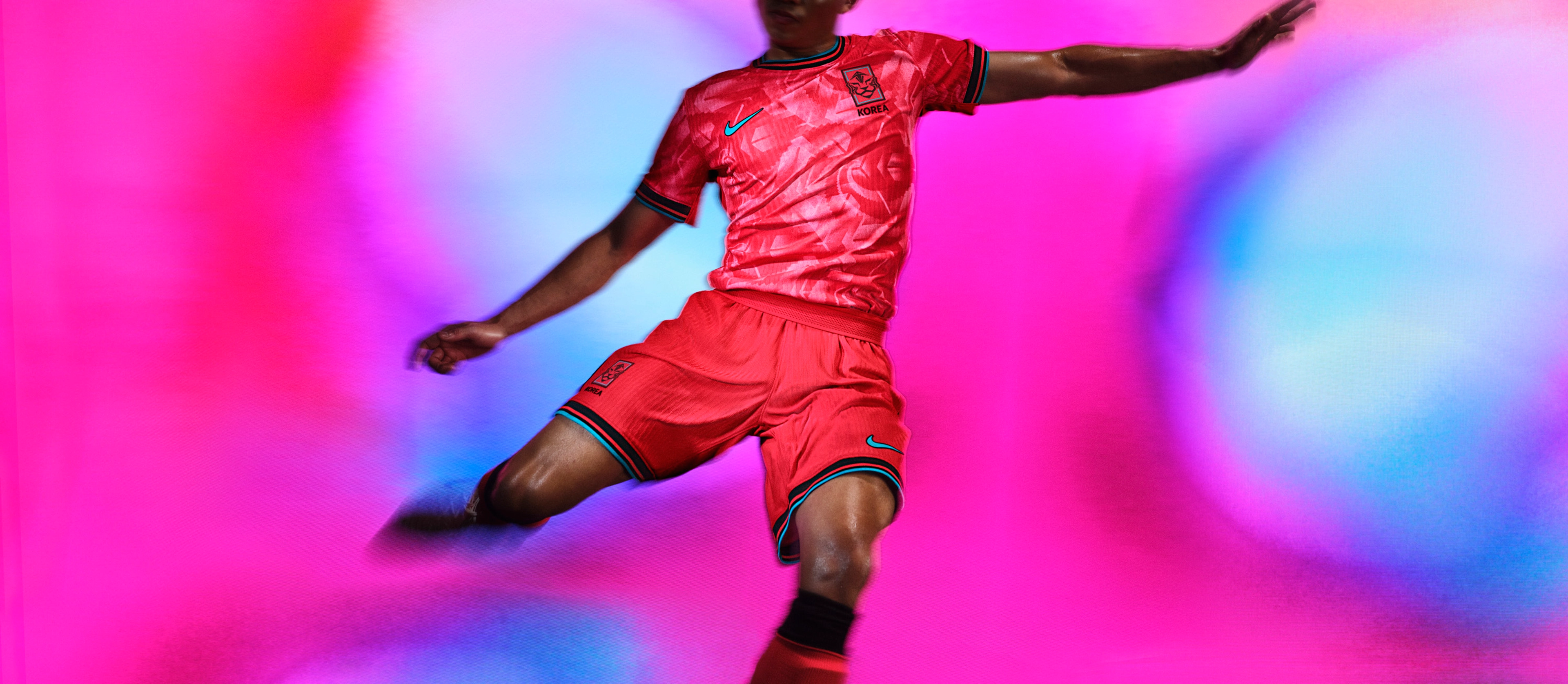 Athlete in a mid-kicking pose wearing a red and pink soccer jersey with black and teal striped trim, as well as and matching red shorts with matching trim, each with a Nike swoosh and team logo on the chest and legs.