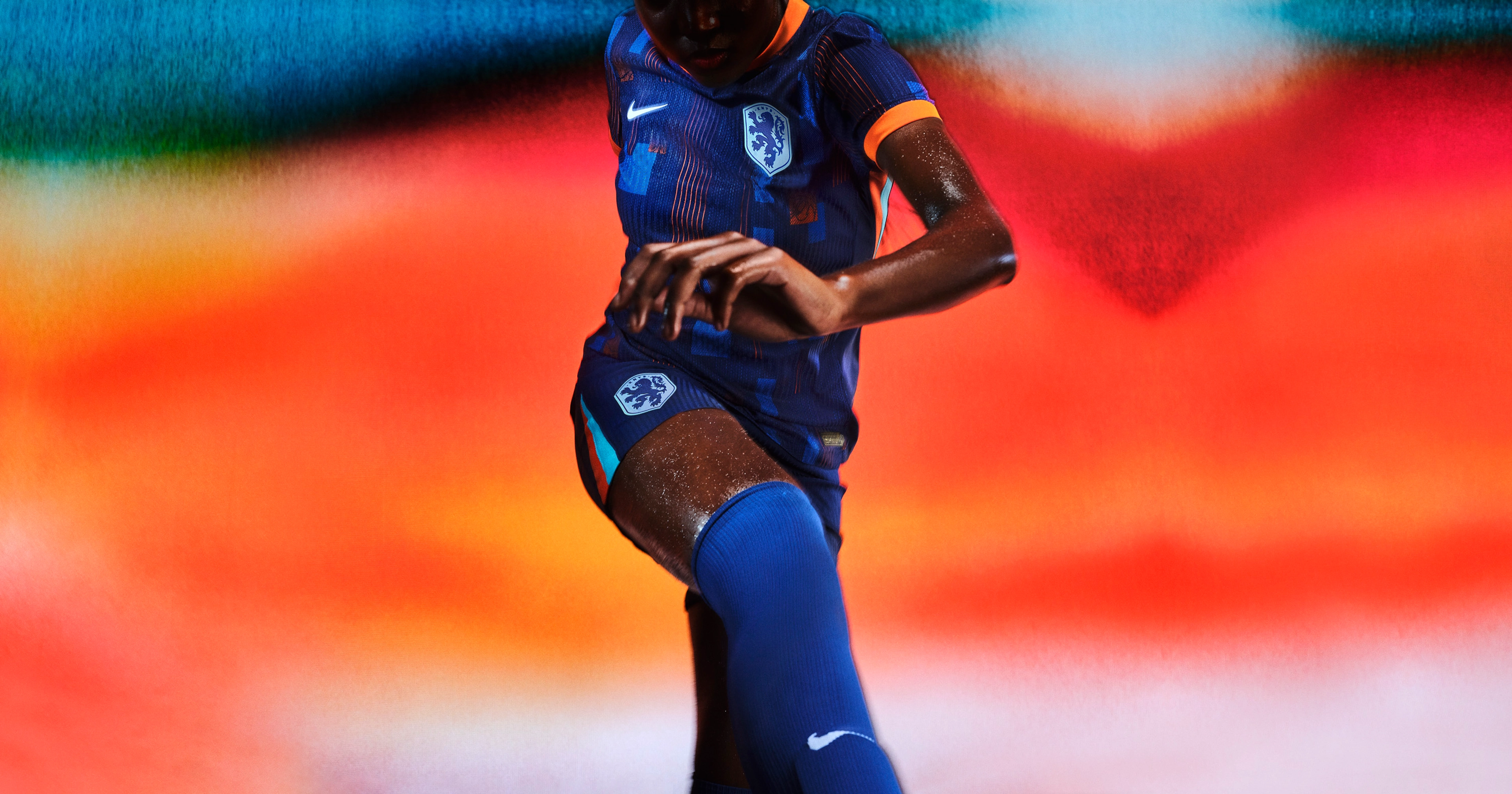Athlete in a kicking pose wearing an indigo soccer jersey with an orange collar and cuffs and blue and orange rectangle patterns on the front, as well as matching indigo blue shorts, each with a Nike swoosh and a team logo on the chest and legs. They are also wearing indigo blue socks.