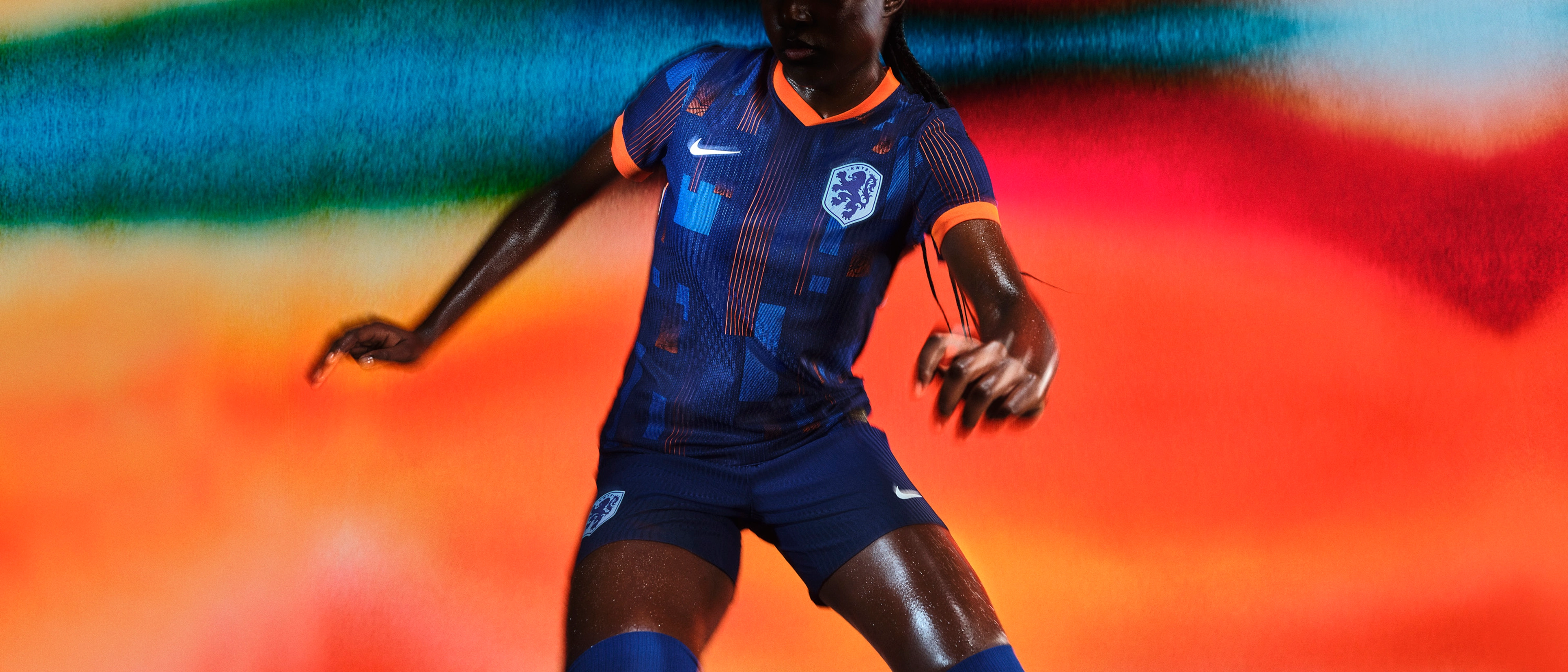 Athlete in a pivoting pose wearing an indigo soccer jersey with an orange collar and cuffs and blue and orange rectangle patterns on the front, as well as matching indigo blue shorts, each with a Nike swoosh and a team logo on the chest and legs.