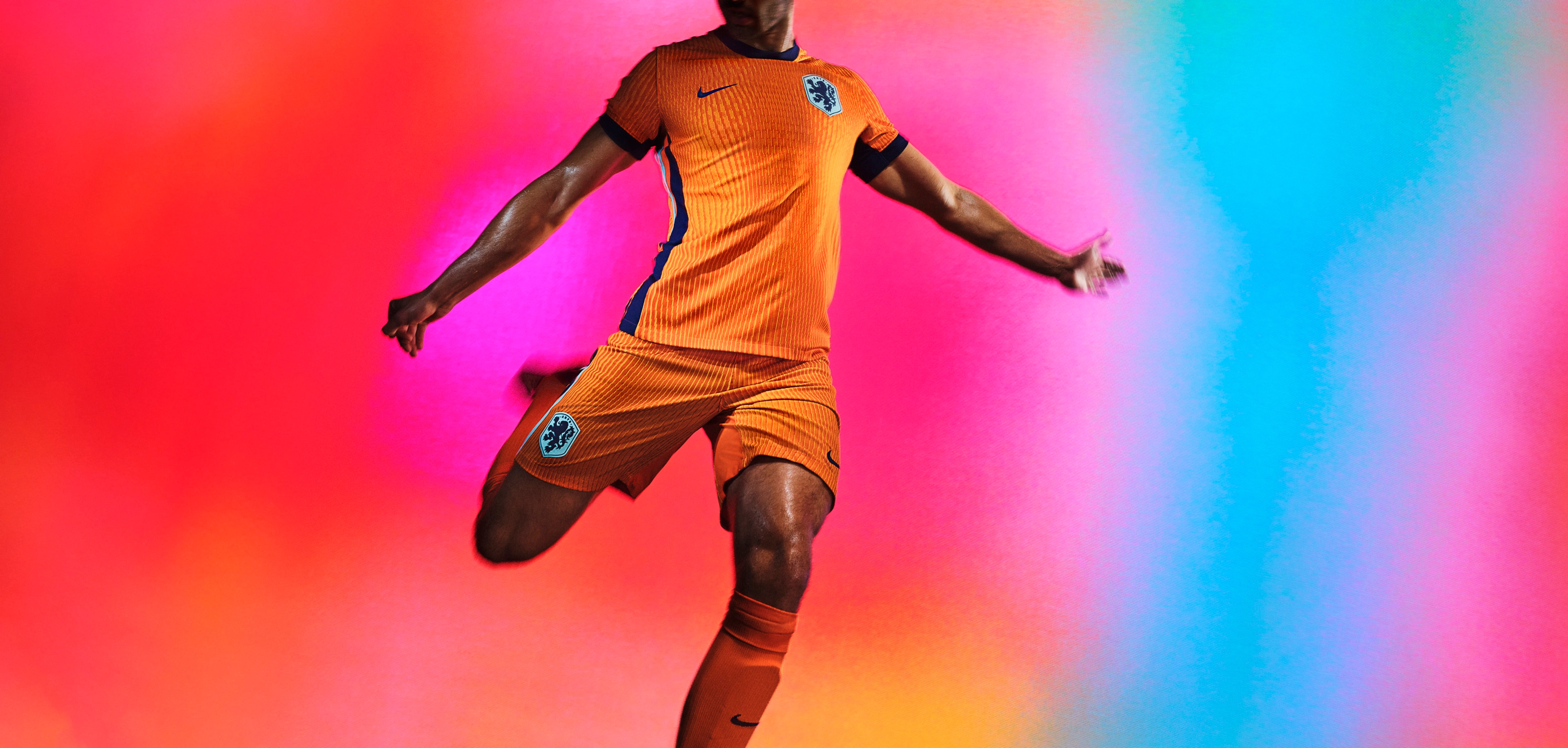 Athlete in a mid-kicking pose wearing an orange soccer jersey with an indigo blue collar, cuffs, and sides, as well as matching orange shorts, each of which have a Nike swoosh and team logo on the chest and legs. They are also wearing matching orange socks.