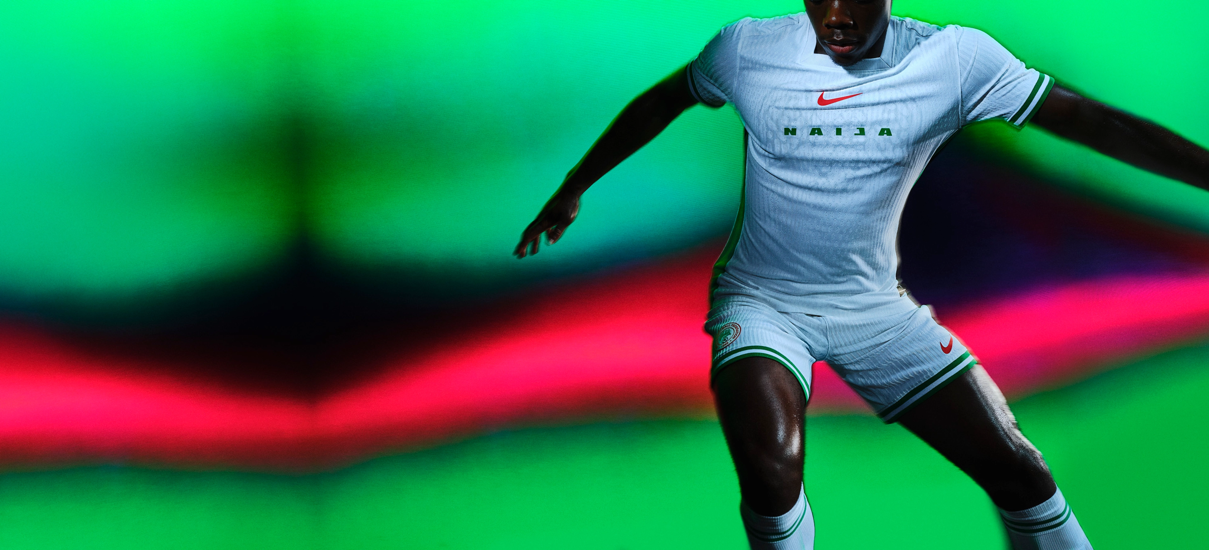 Athlete dribbling a soccer ball wearing a white soccer jersey with green stripes on the cuffs and sides, as well as matching white shorts with green trim, each with a Nike swoosh and team logo on the chest and legs. They are also wearing white socks with matching green striped trim.