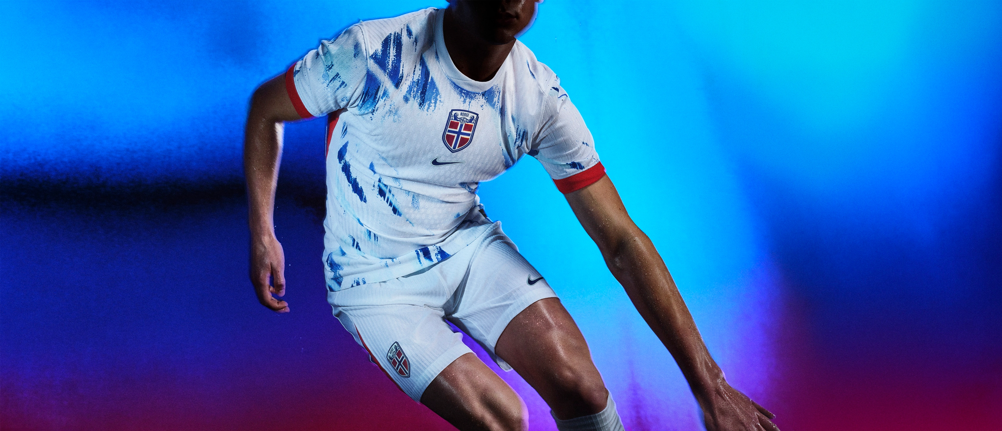 Athlete in a running pose wearing a white soccer jersey with blue brushstroke patterns, red sides, and red cuffs, as well as matching white shorts, each with a Nike swoosh and team logo on the chest and legs.