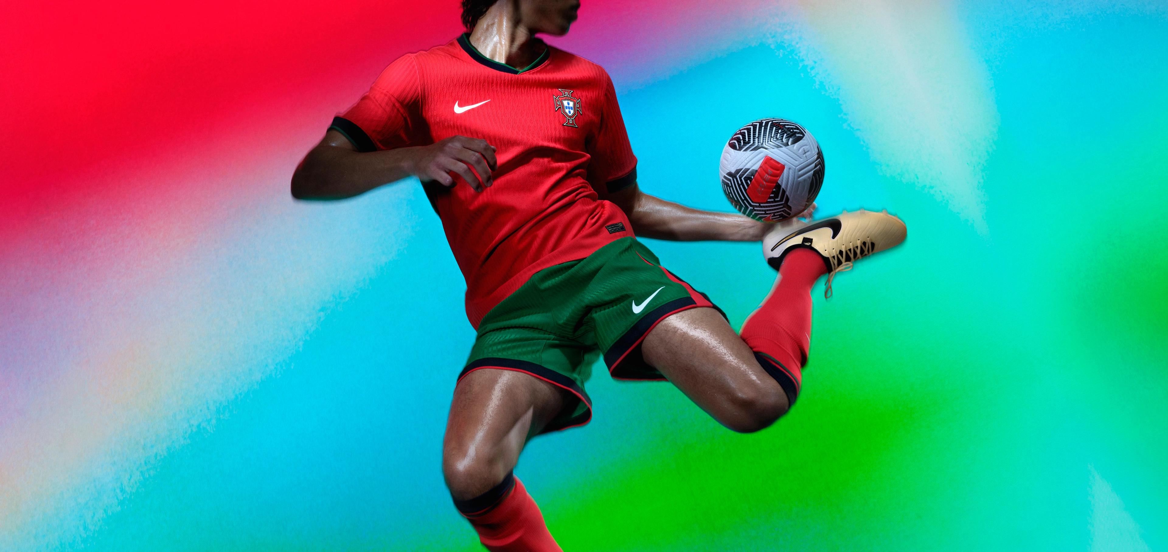 Athlete bouncing a soccer ball off their heel and wearing a red jersey with a forest green collar and forest green cuffs, as well as matching forest green shorts with red trim with a Nike swoosh and team logo on the chest and legs. They are also wearing matching red socks with green trim.