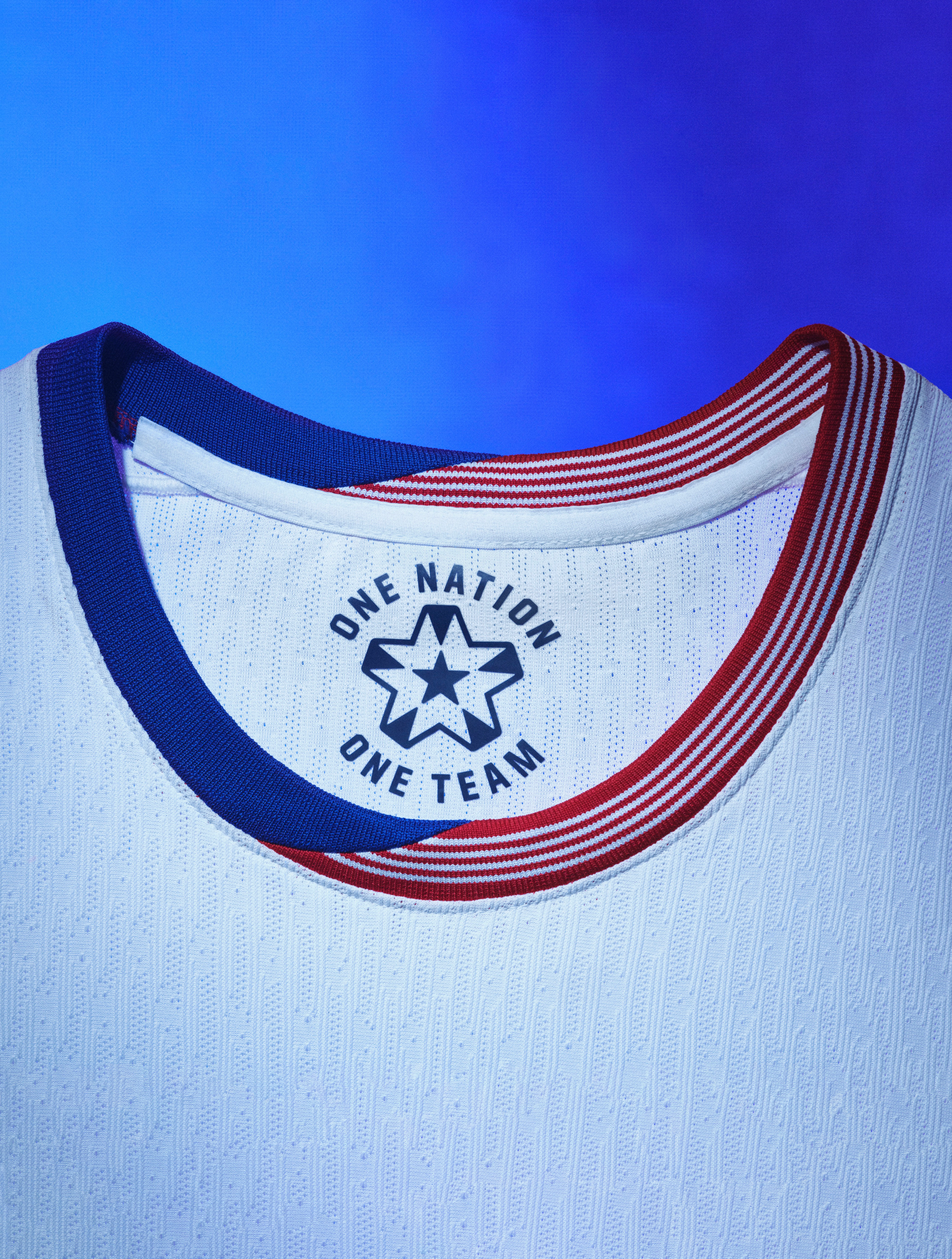 Close up view of a white soccer jersey's collar with blue trim coupled with red and white striped trim and a logo on the back.