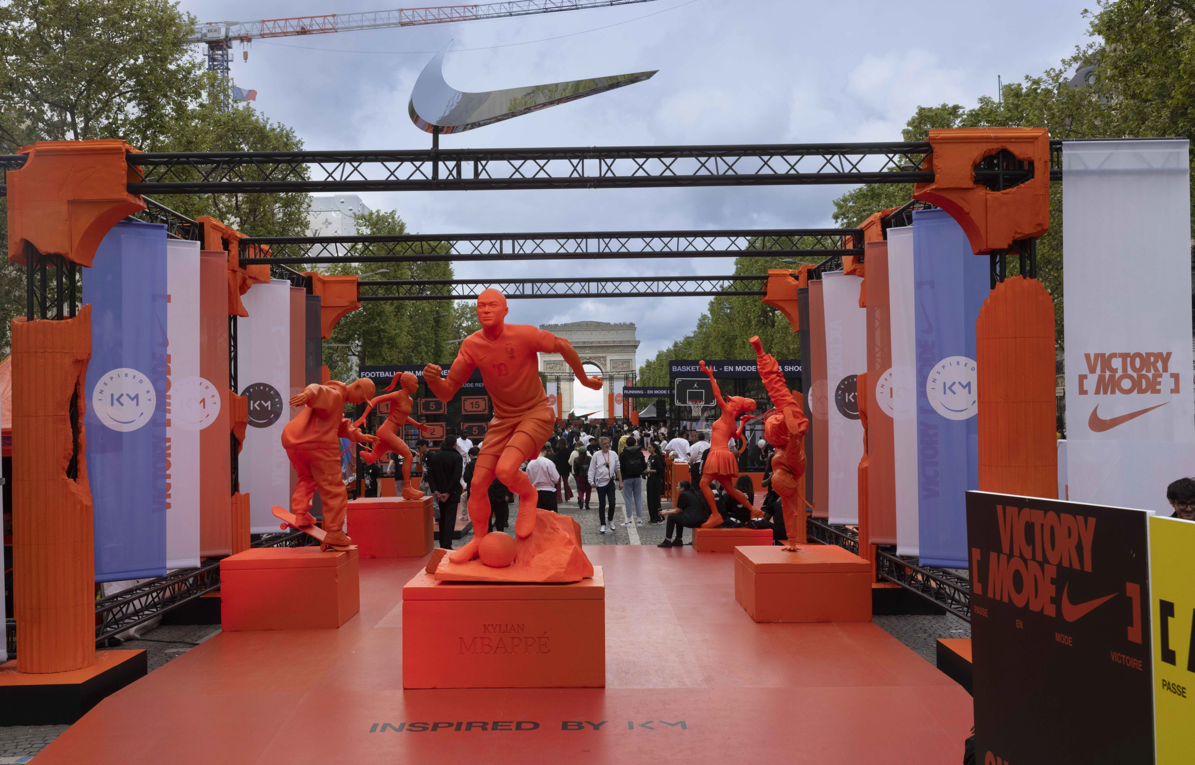 Front view of a pavilion of bright orange statues depicting athletes doing different sports, at the Victory Mode event in Paris, France. Statues are surrounded by orange pillars and blue, white, and orange banners.