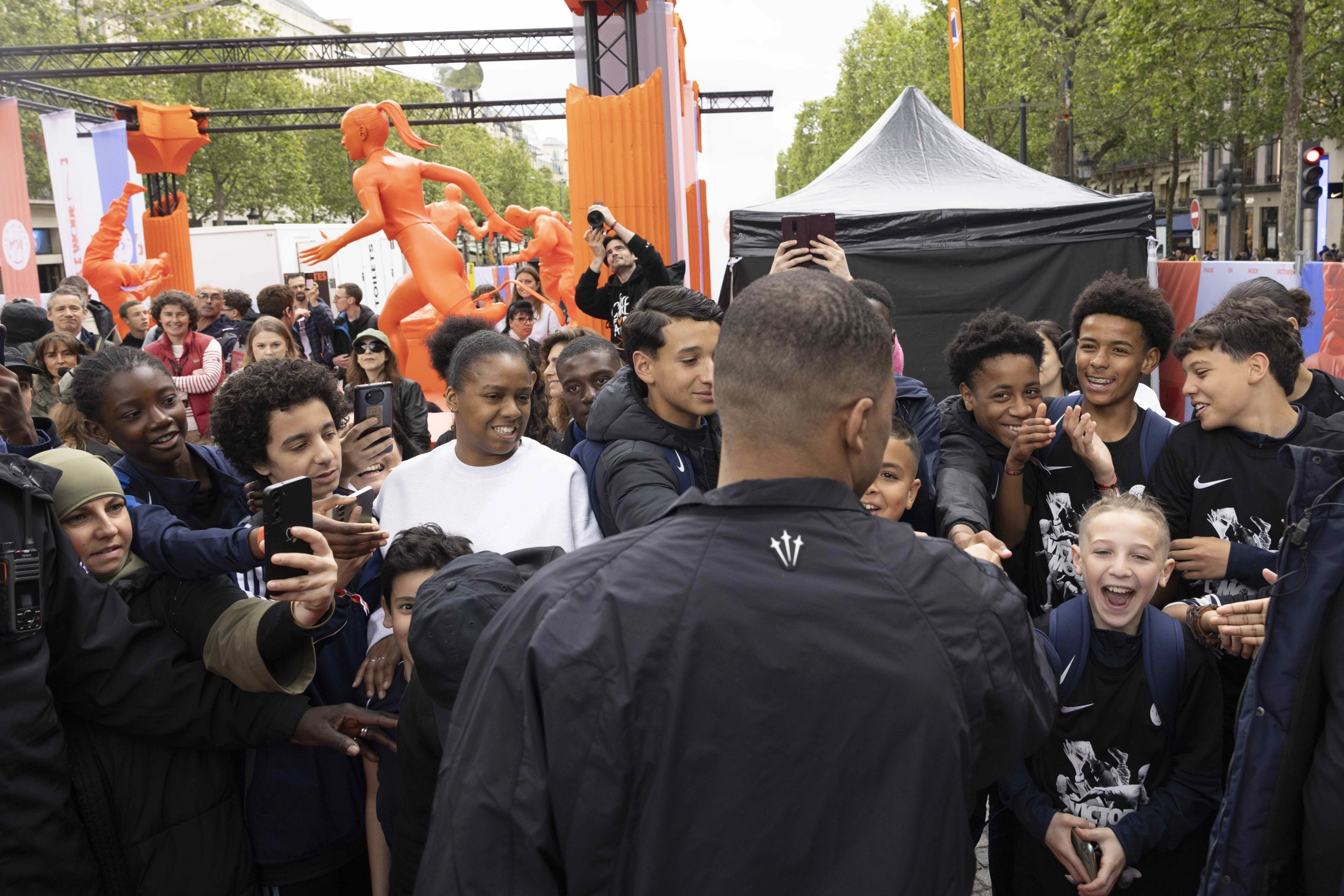 Back view of Kylian Mbappe, as he faces a crowd of kids at the Victory Mode event in Paris, France. The energized crowd is smiling and capturing photos of Mbappe. In the background are bright orange statues of kids doing sport.