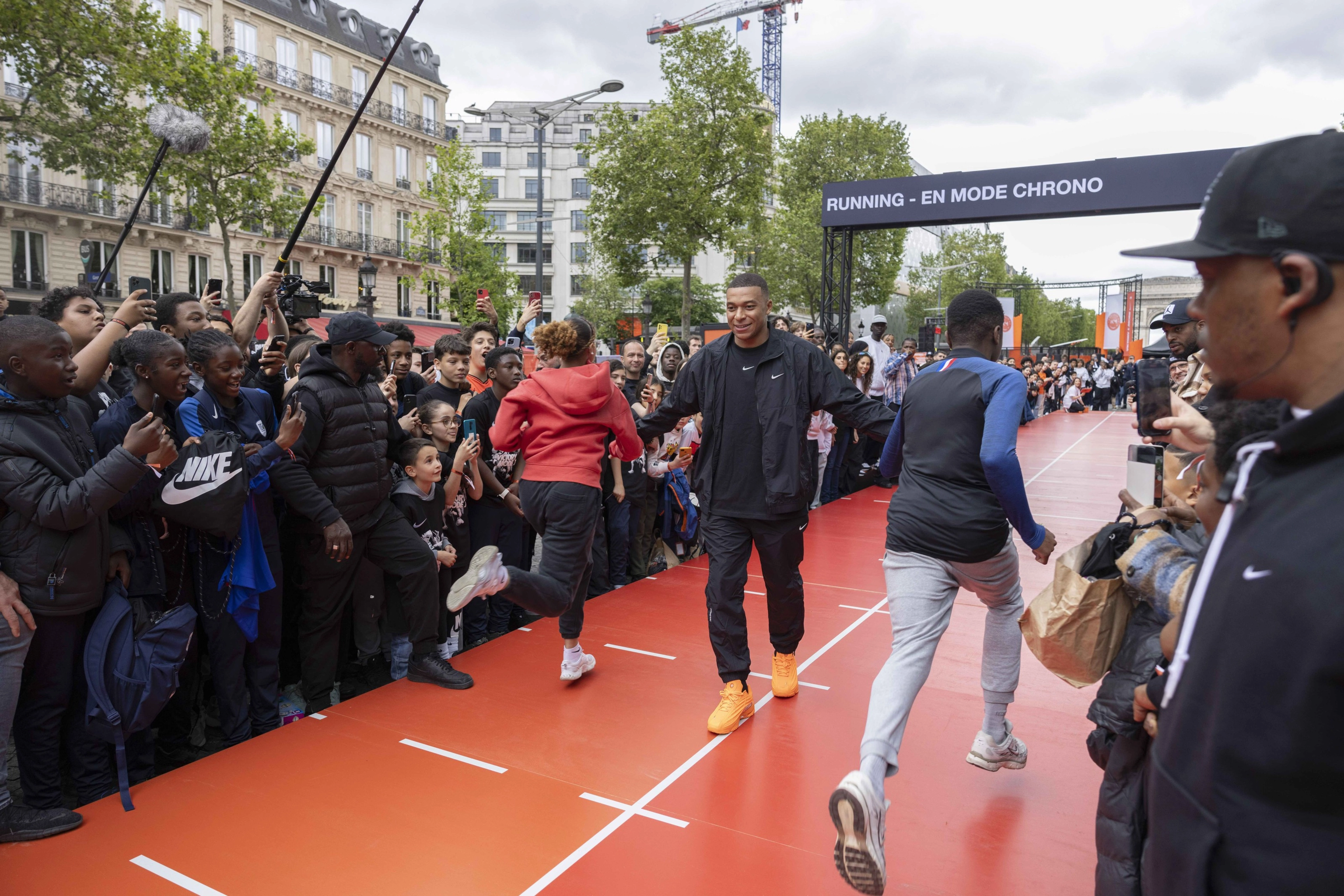 Front view of Kylian Mbappe wearing a black Nike tee, jacket, and pants, while walking between 2 runners on an orange stage at the Victory Mode event in Paris, France. A crowd surrounds the activity on the stage.