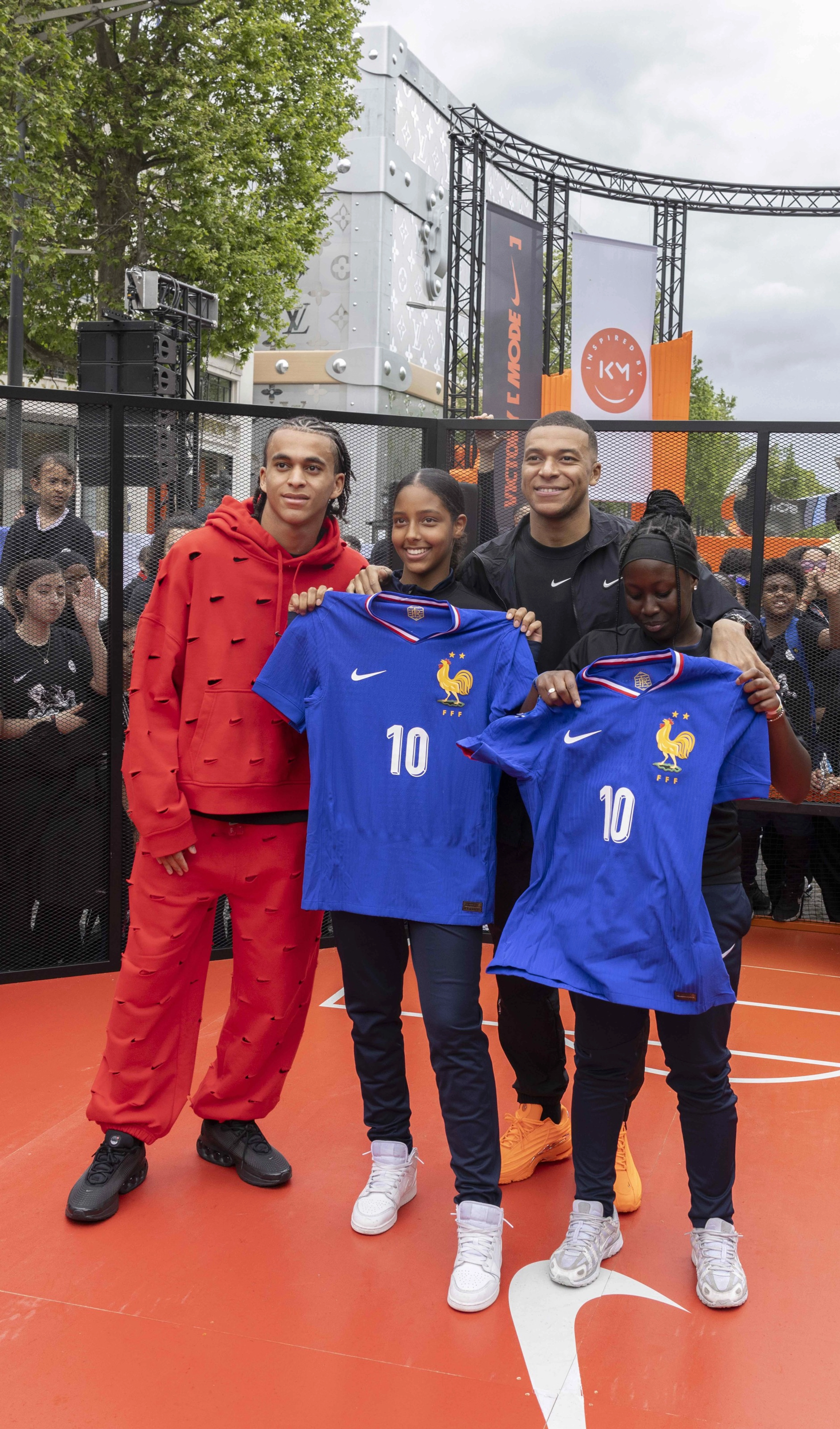 Kylian and Ethan Mbappe pose with two girls, who are holding #10 French World Cup jerseys in bright blue. They are standing on an orange court in front of the House of Innovation in Paris at the Victory Mode event.