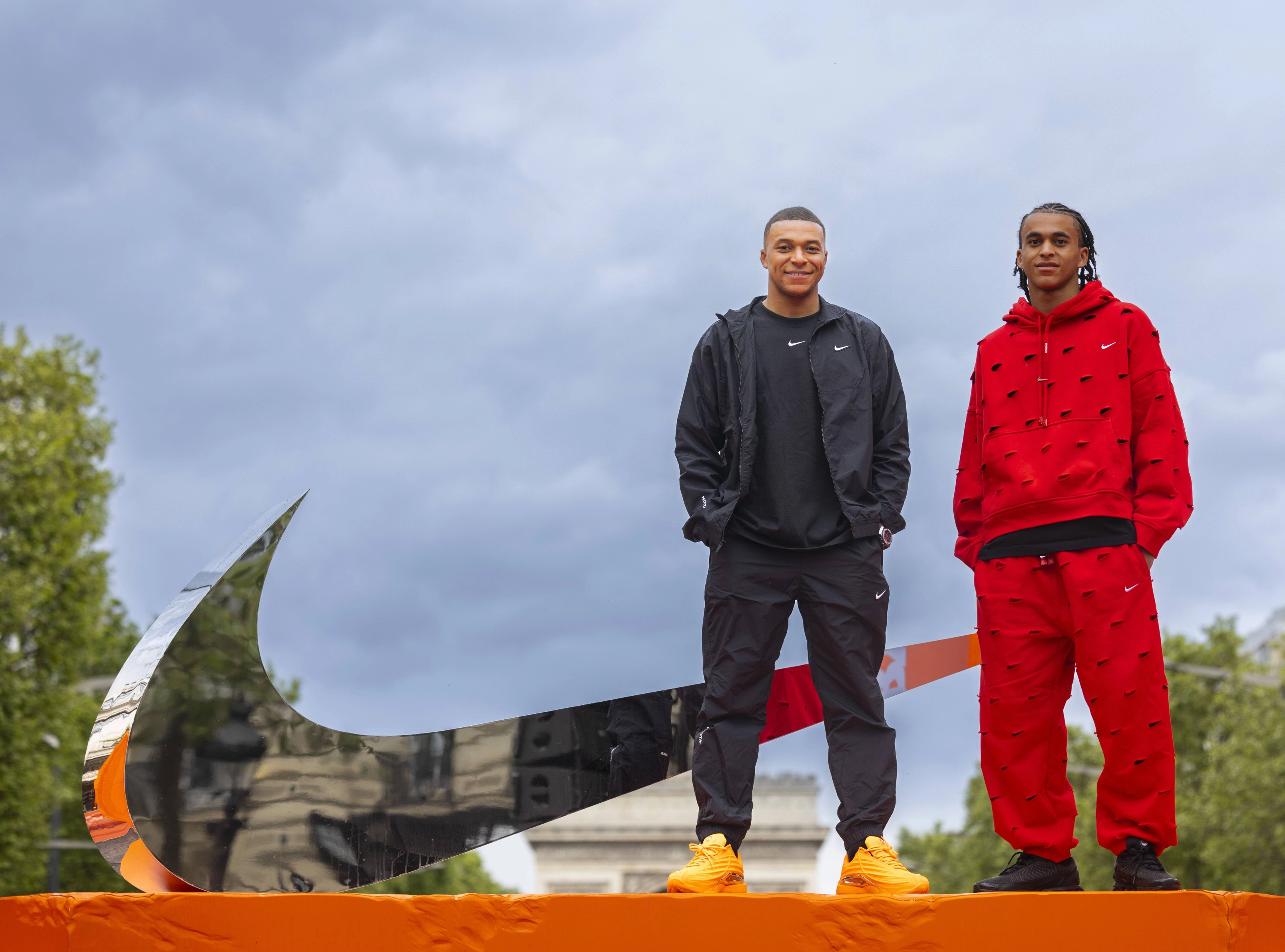 Front view of Kylian Mbappe, and his brother, Ethan Mbappe, as they stand next to a large mirrored Swoosh. Kylian wears black Nike tee, jacket and pants with orange footwear.  Ethan wears a red fleece hoodie and pants with black footwear. Both smile towards the camera.