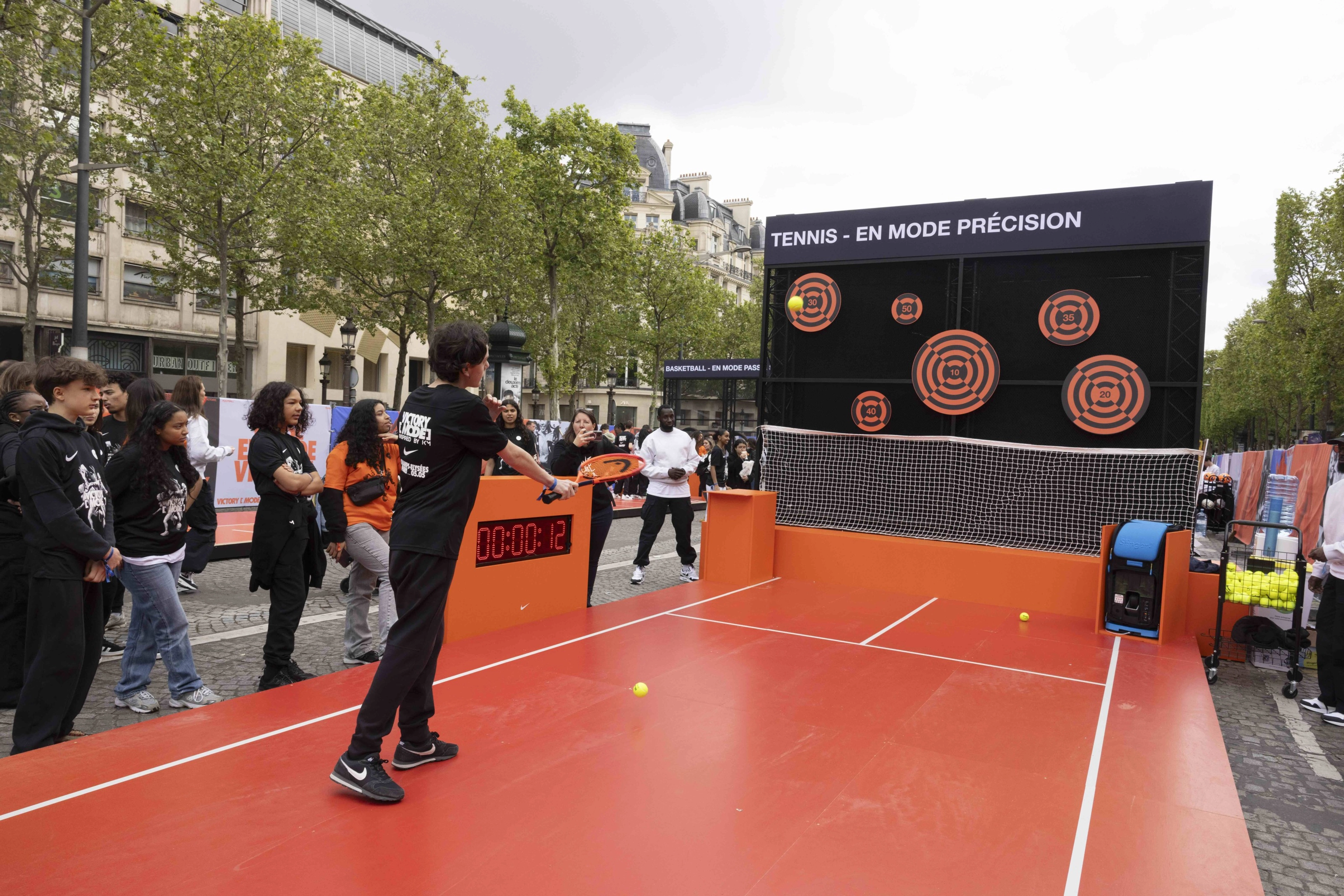 Back view of a young athlete wearing a black Victory Mode tee using a bright orange tennis racket to participate in the Tennis Challenge at the Victory Mode event in Paris, France.  A crowd looks on as he aims at orange and black targets.