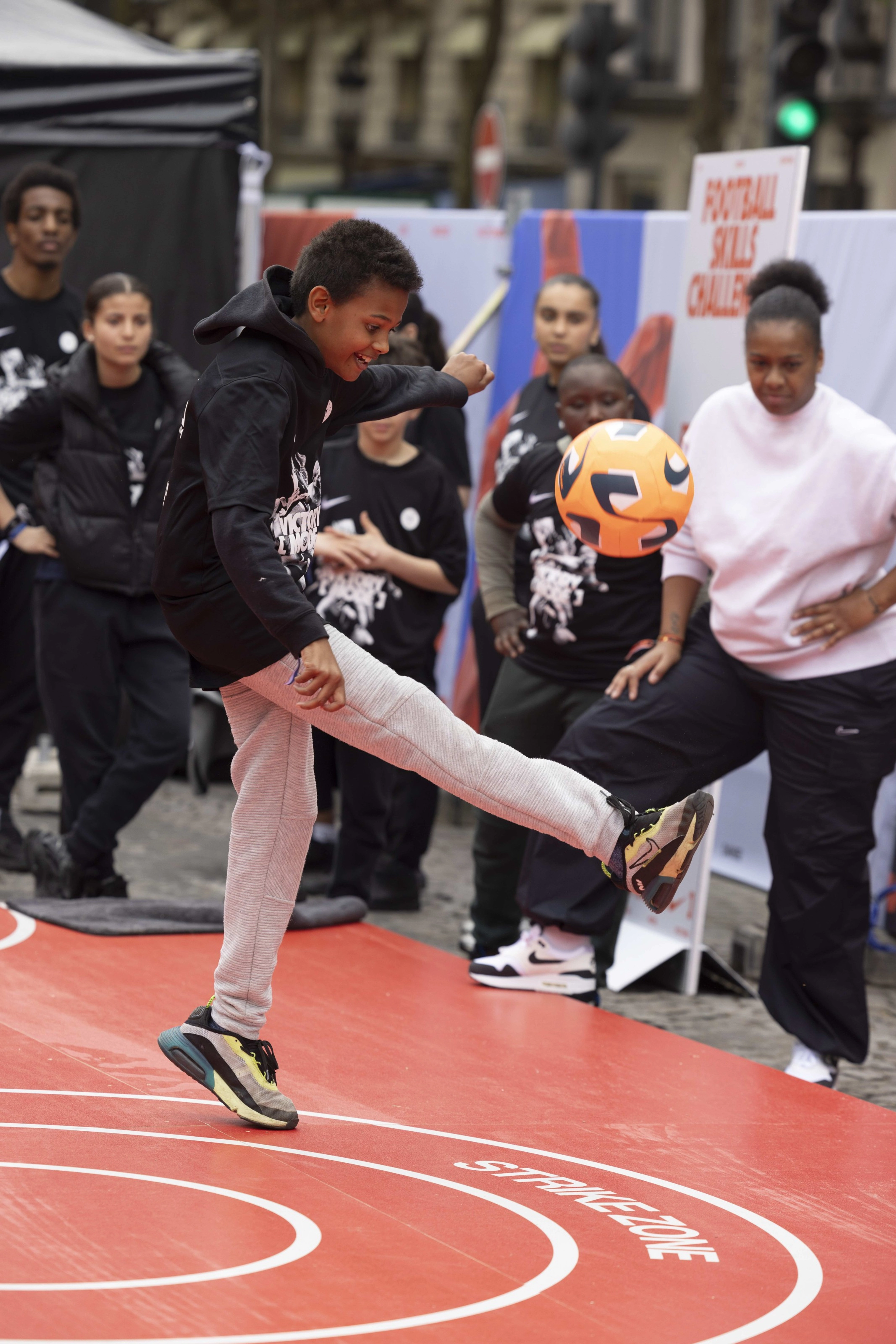 A side view of a young boy juggling a bright orange football on a red surface that has a strikezone grapic printed on it. The boy has a big smile as a crowd looks on.