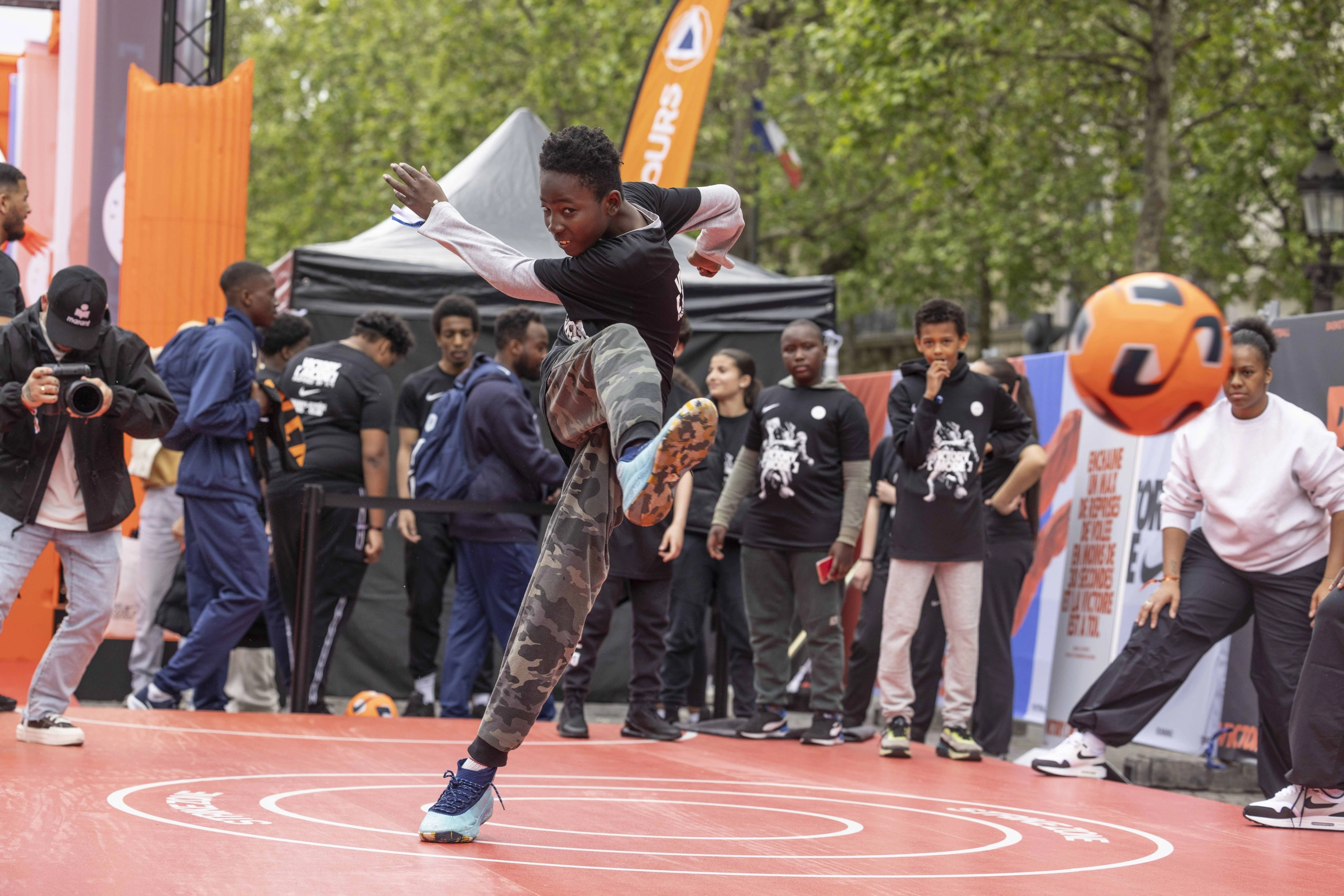Front view of a young athlete kicking a bright orange soccer ball with great energy. His body twists as he strikes the ball in front of a crowd of onlookers.