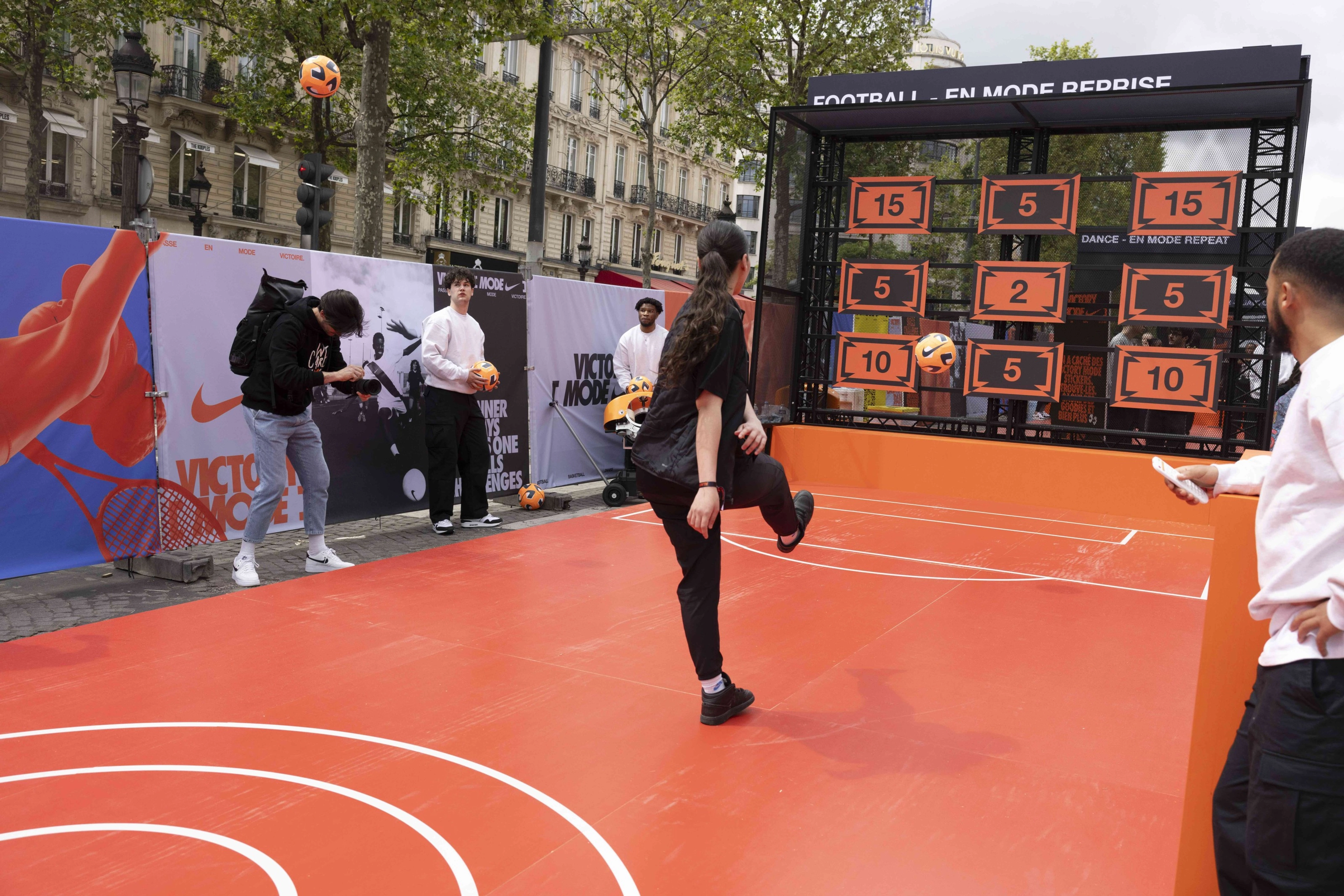 Back view of a young athlete kick an orange football during a football challenge at the Victory Mode event in Paris, France.  She stands on a bright orange court, with photographers and event helpers nearby.