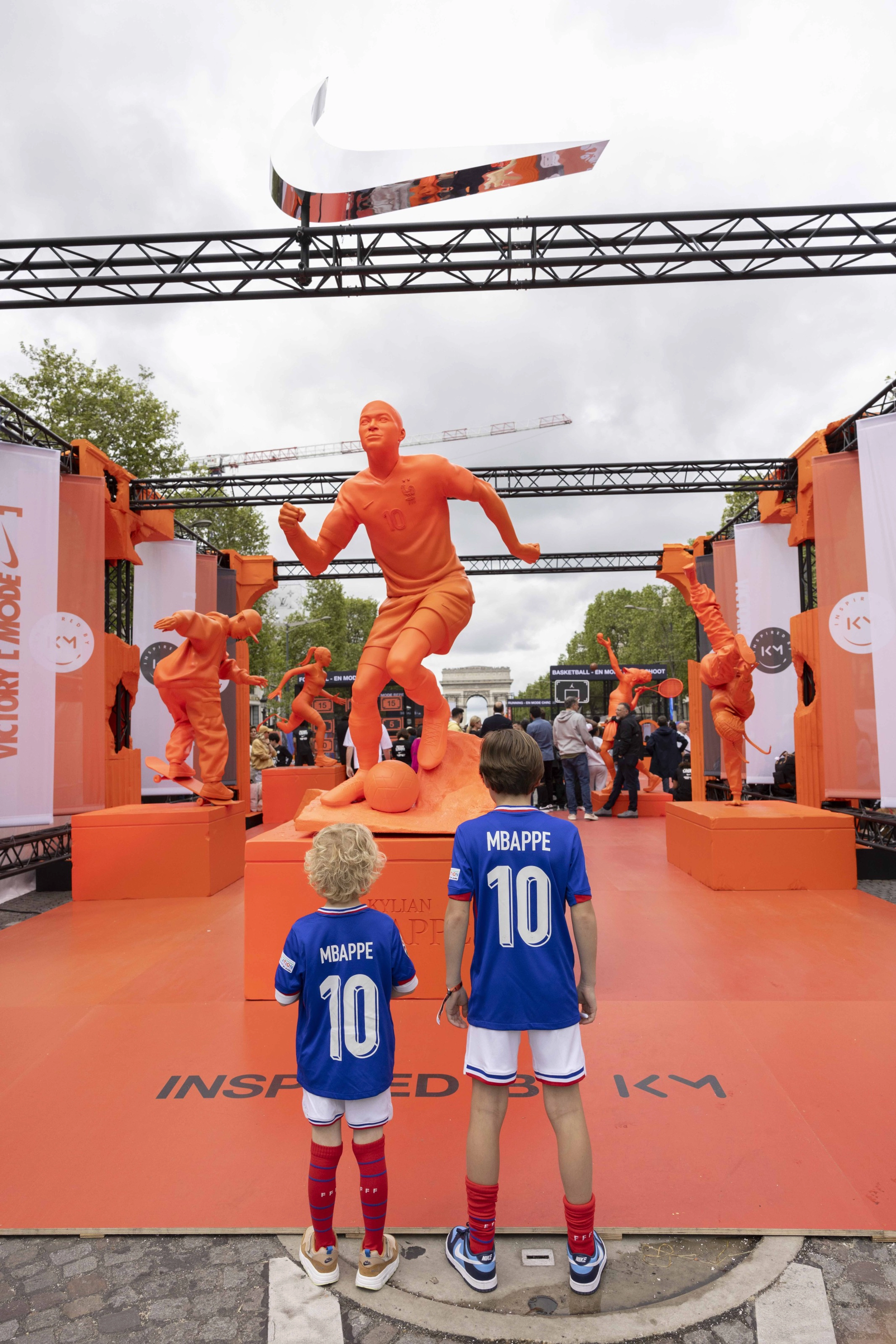 Back view of two young boys dressed in French football kits, including bright blue #10 jerseys, looking at a bright orange statue of Kylian Mbappe dribbling a football.