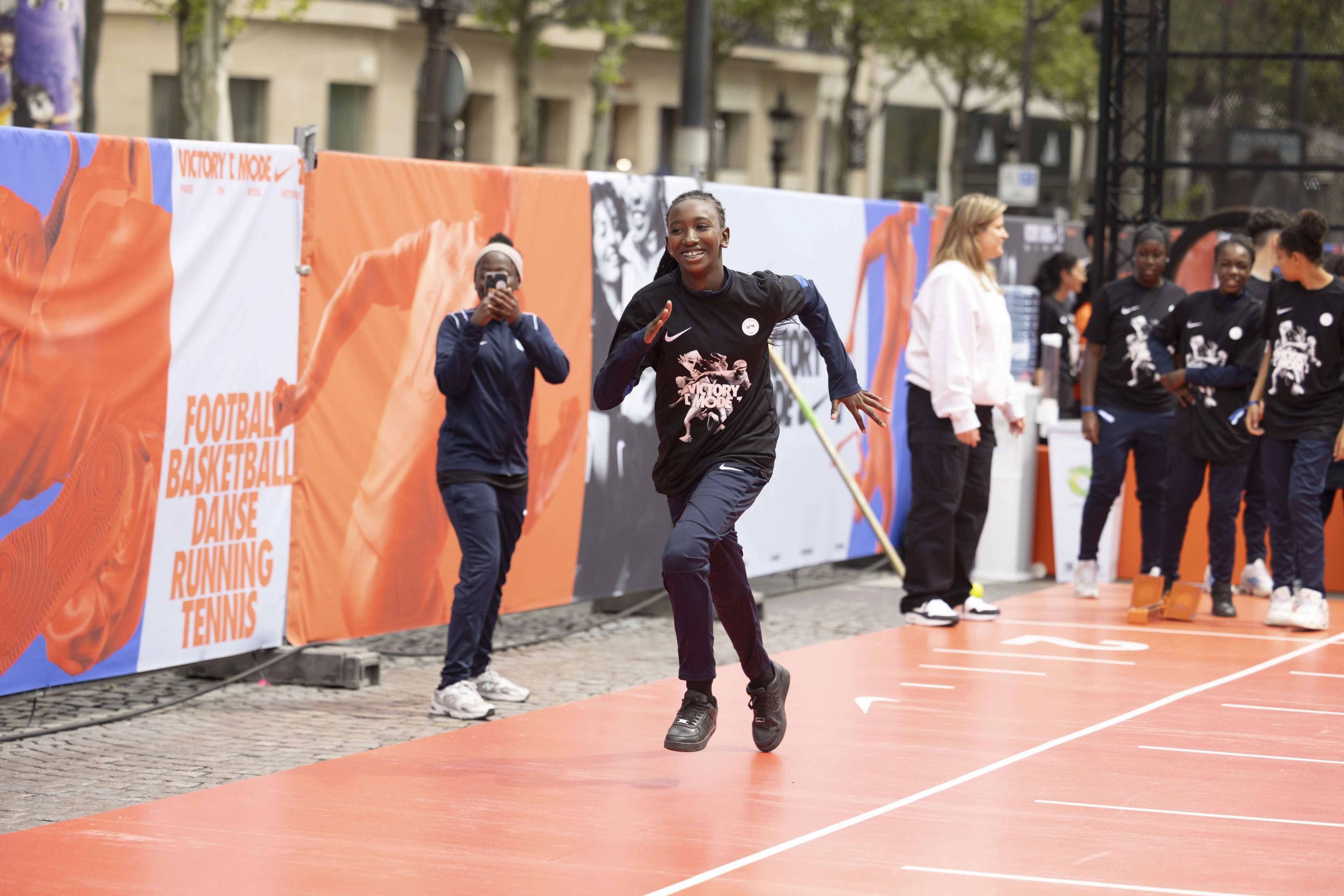Front angled view of a girl in a black Victory Mode event tee shirt running down the orange track, as another young athlete snaps a picture on her phone. There is a crowd of kids in the background, waiting to run.