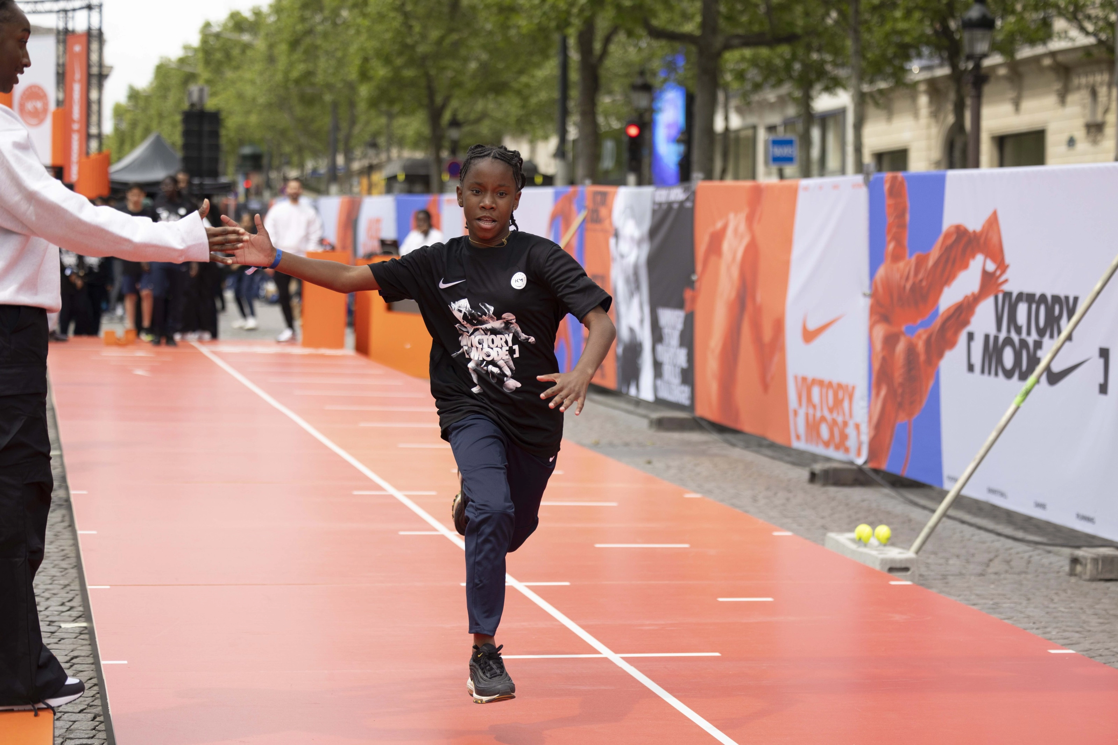 Front view of a girl in a black Victory Mode event tee shirt running down the orange track, receiving a high five from an onlooker.