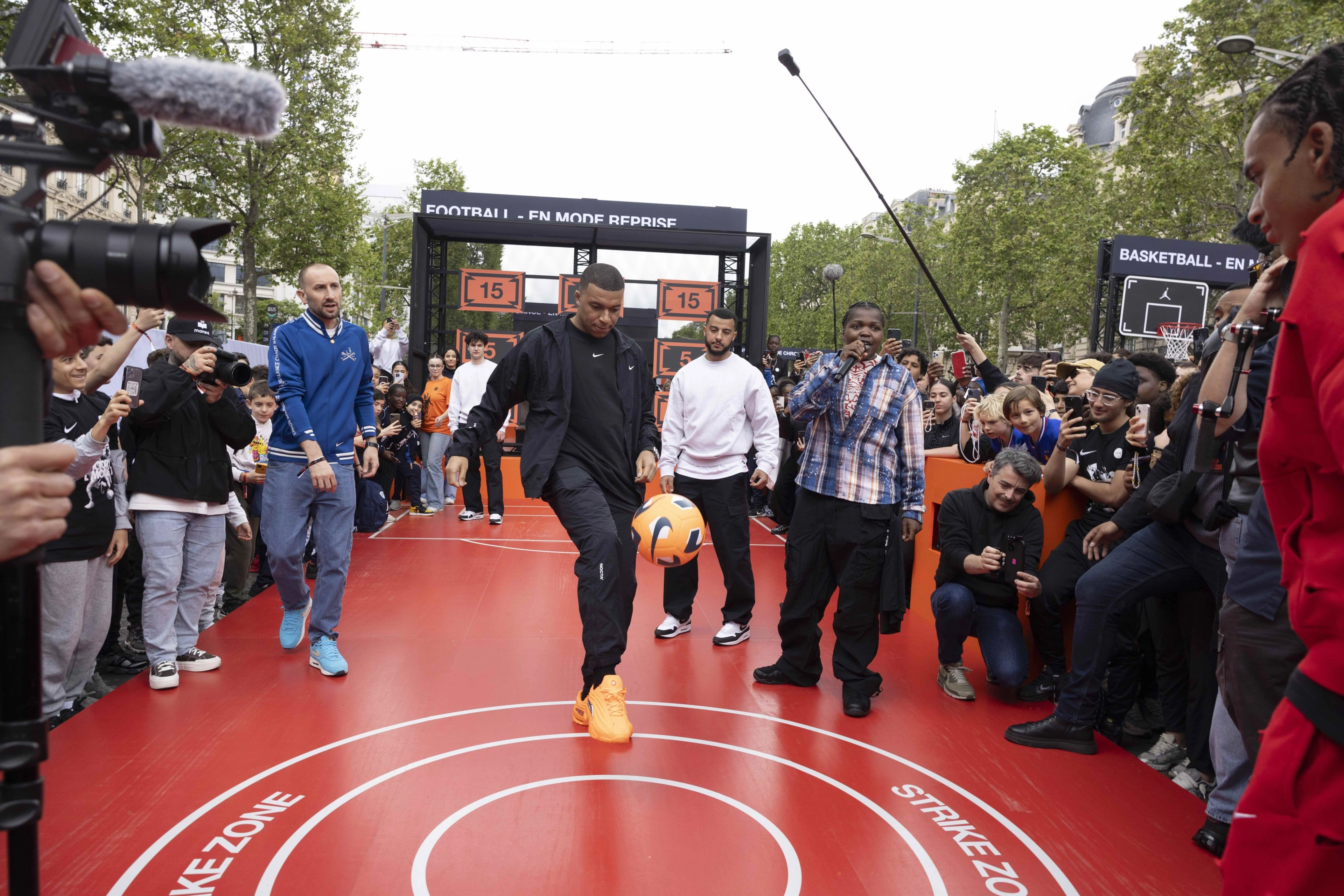 Front view of Kylian Mbappe juggling an orange Nike football on a red platform in front of a crowd of young athletes and photographers at the Victory Mode event in Paris, France.