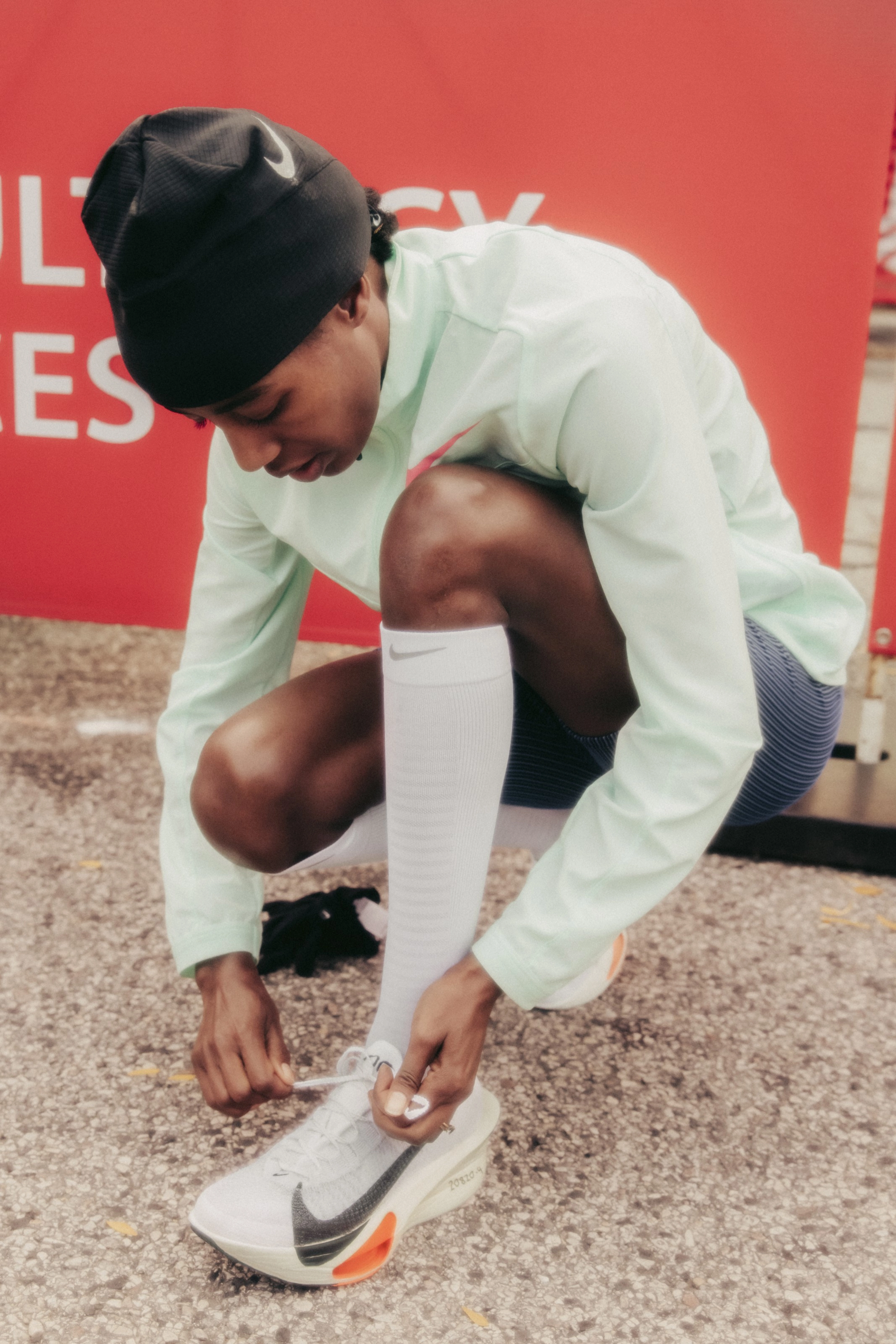 Sifan Hassan bends forward to tied her Nike running shoe pre race. She wears a black beanie, a longsleeve warmup top, and high white socks.