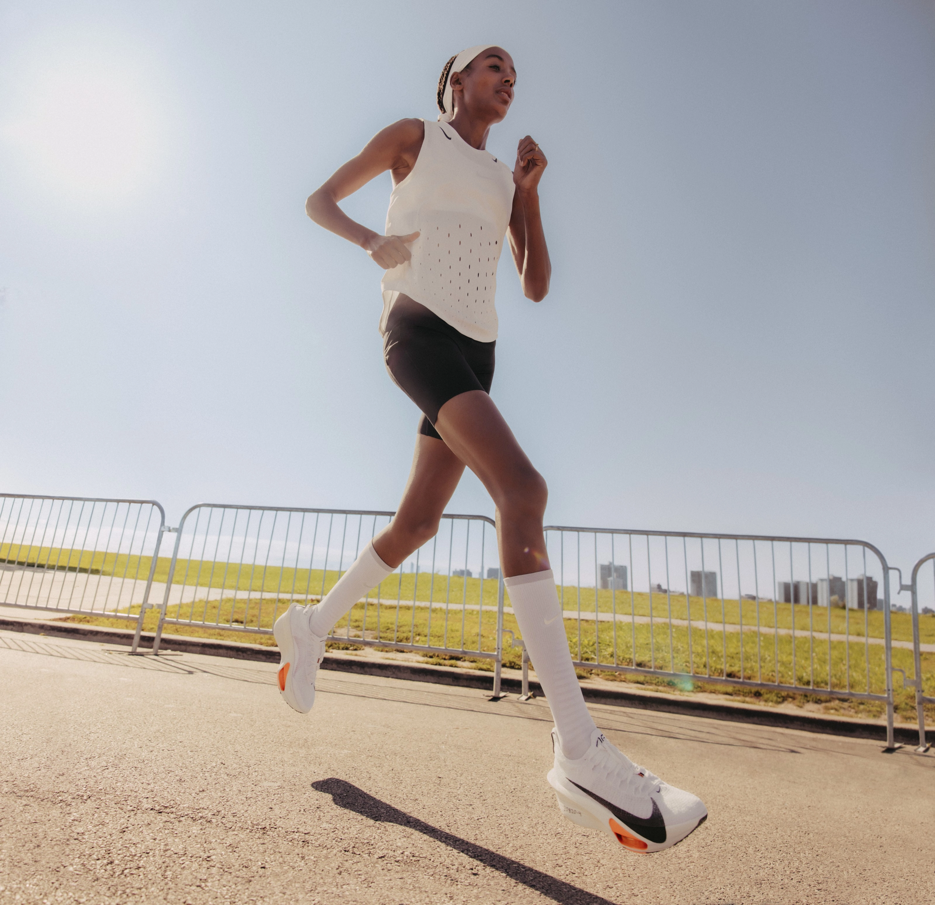 Full body view of Sifan Hissan running on a road. She is in a white Nike running top, blue shorts, Nike running shoes, and high white socks. Barriers edge the road and there is a city silhouette in the background.