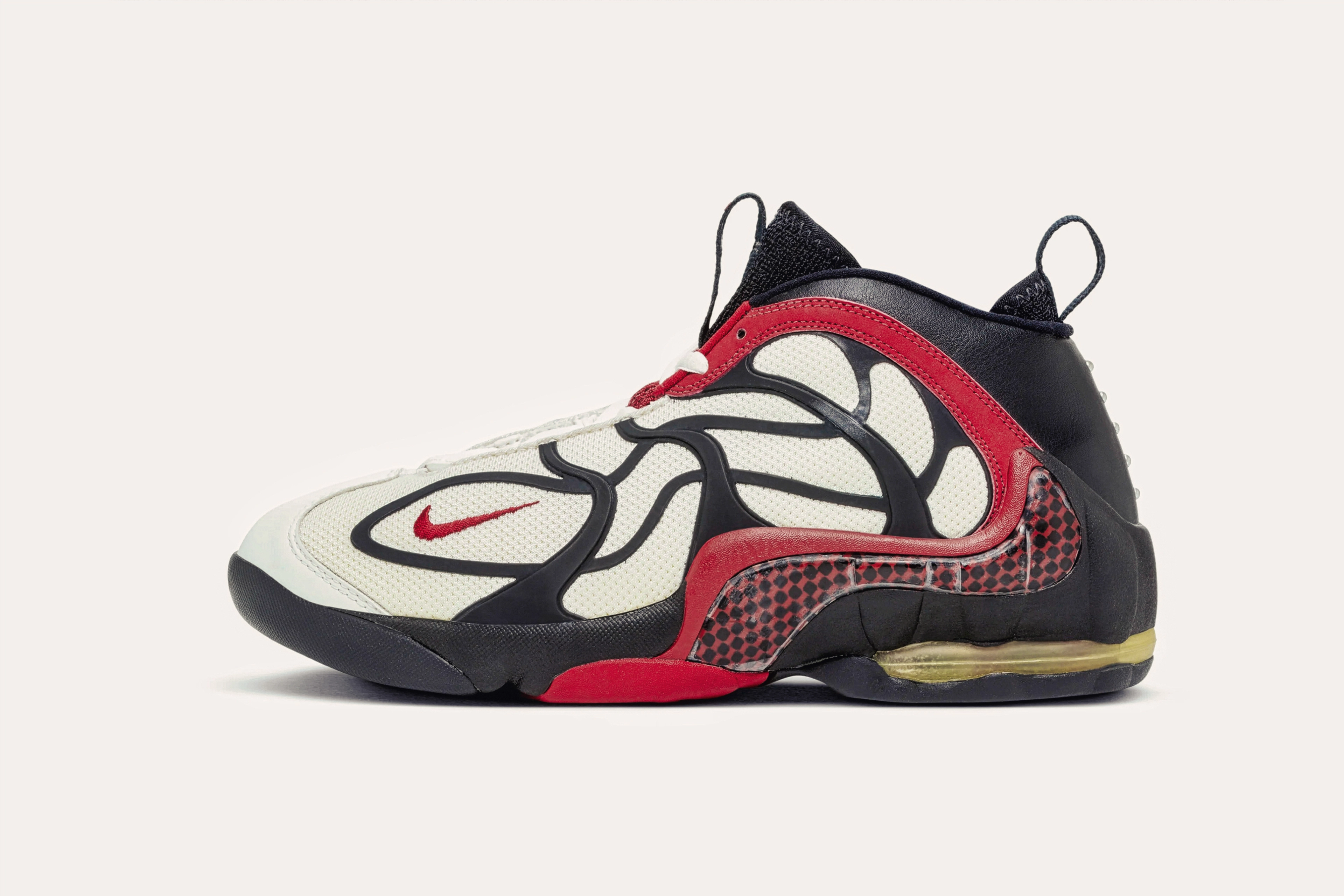 A white sneaker with a black leather backing and a black web-like pattern with red trim.