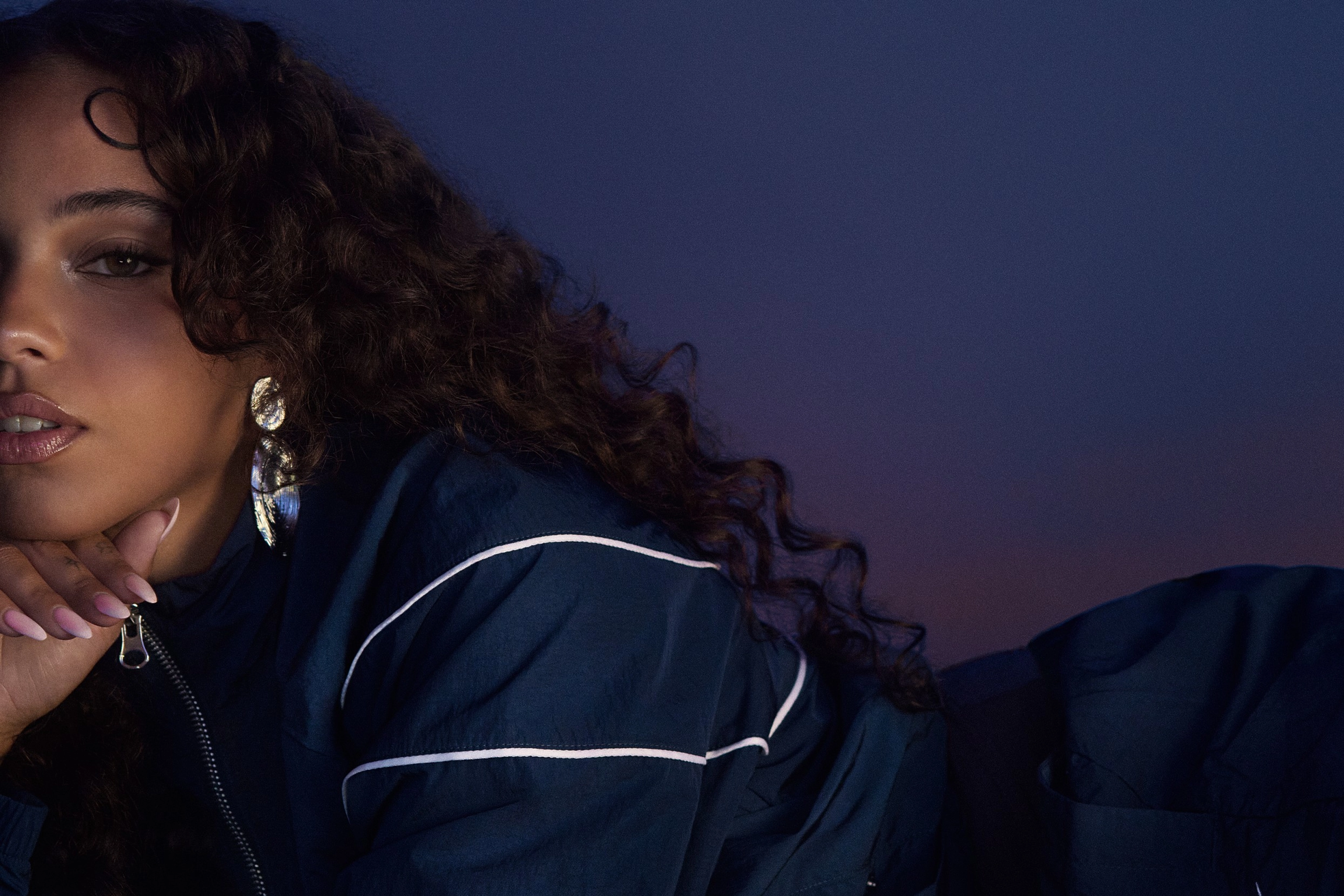 Maria Isabel lies against a blue/purple gradient background, wearing a navy blue Nike Windrunner tracksuit. We see part of her body and face, as she props her hand under her chin, and looks directly at the camera. She wears silver earrings that reflect the blue/purple light.