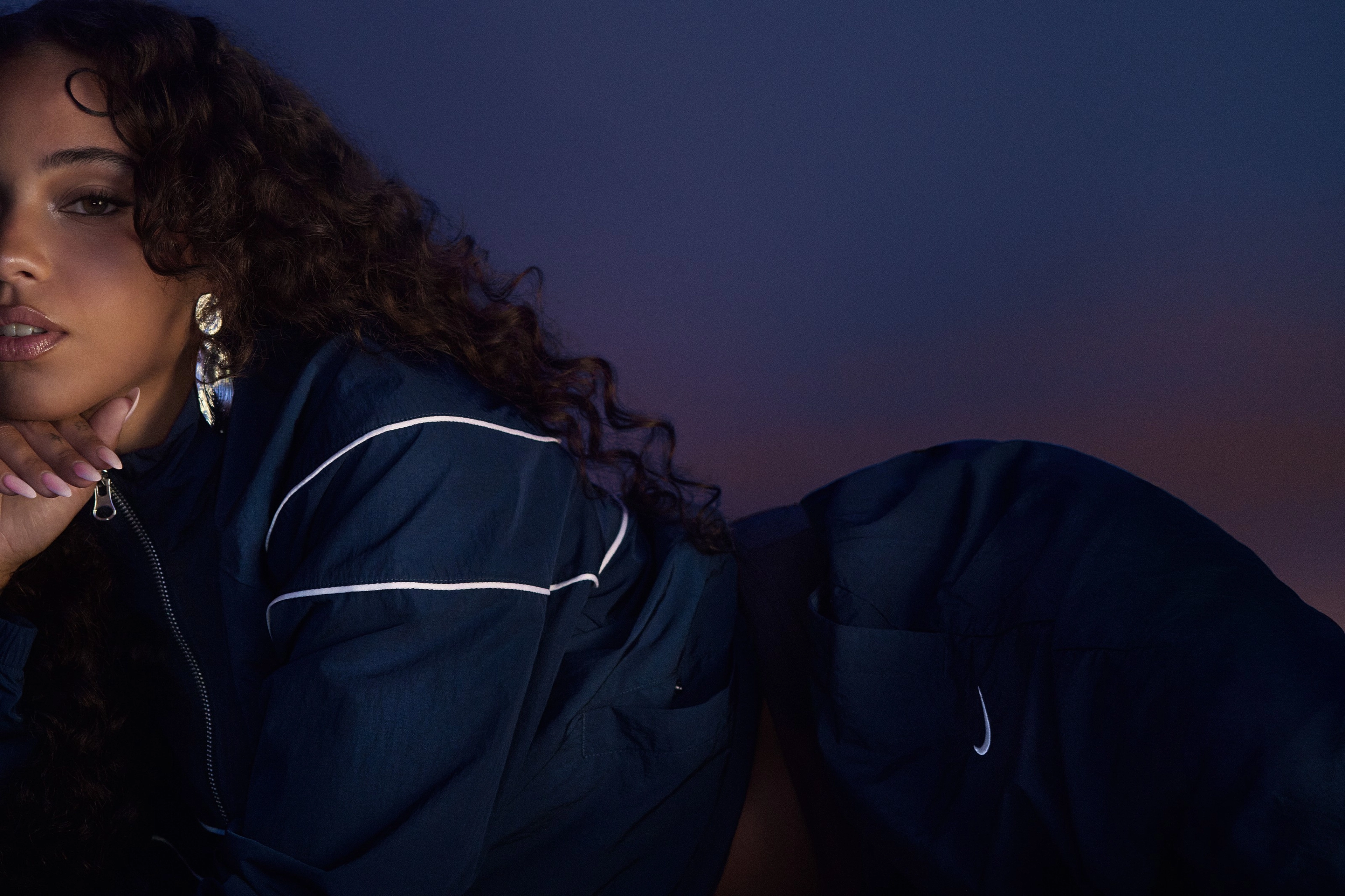 Maria Isabel lies against a blue/purple gradient background, wearing a navy blue Nike Windrunner tracksuit. We see part of her body and face, as she props her hand under her chin, and looks directly at the camera. She wears silver earrings that reflect the blue/purple light.