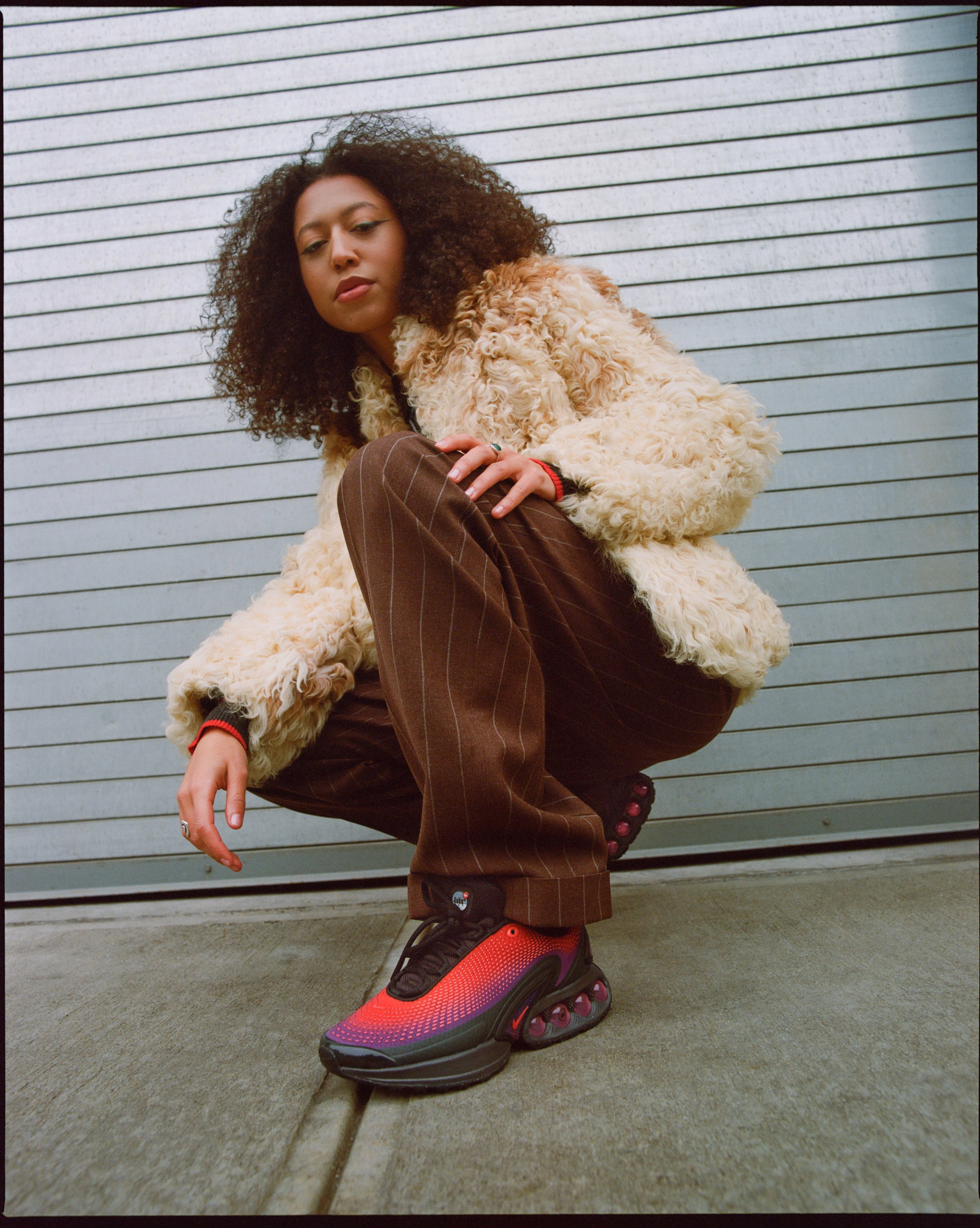 A female model wearing the Nike Air Max Dn in black and red while in a in a squatting position outside on the sidewalk.