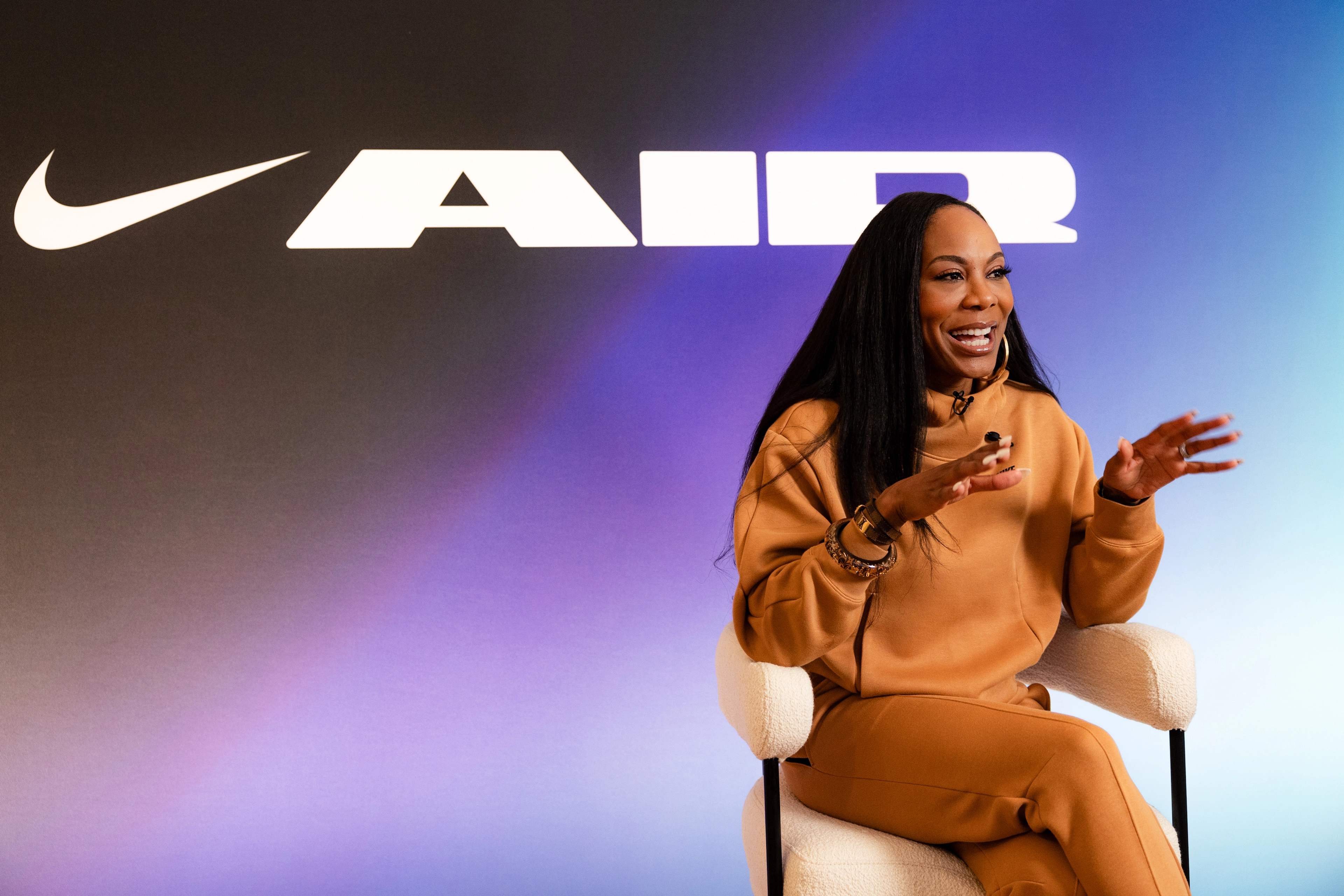 Front angled view of Sanya Richards-Ross wearing Nike Sportswear Tech Fleece in the Flax color. Sanya sits on a white stool, against a blue & purple gradient background with a large white logo for Nike AIR.