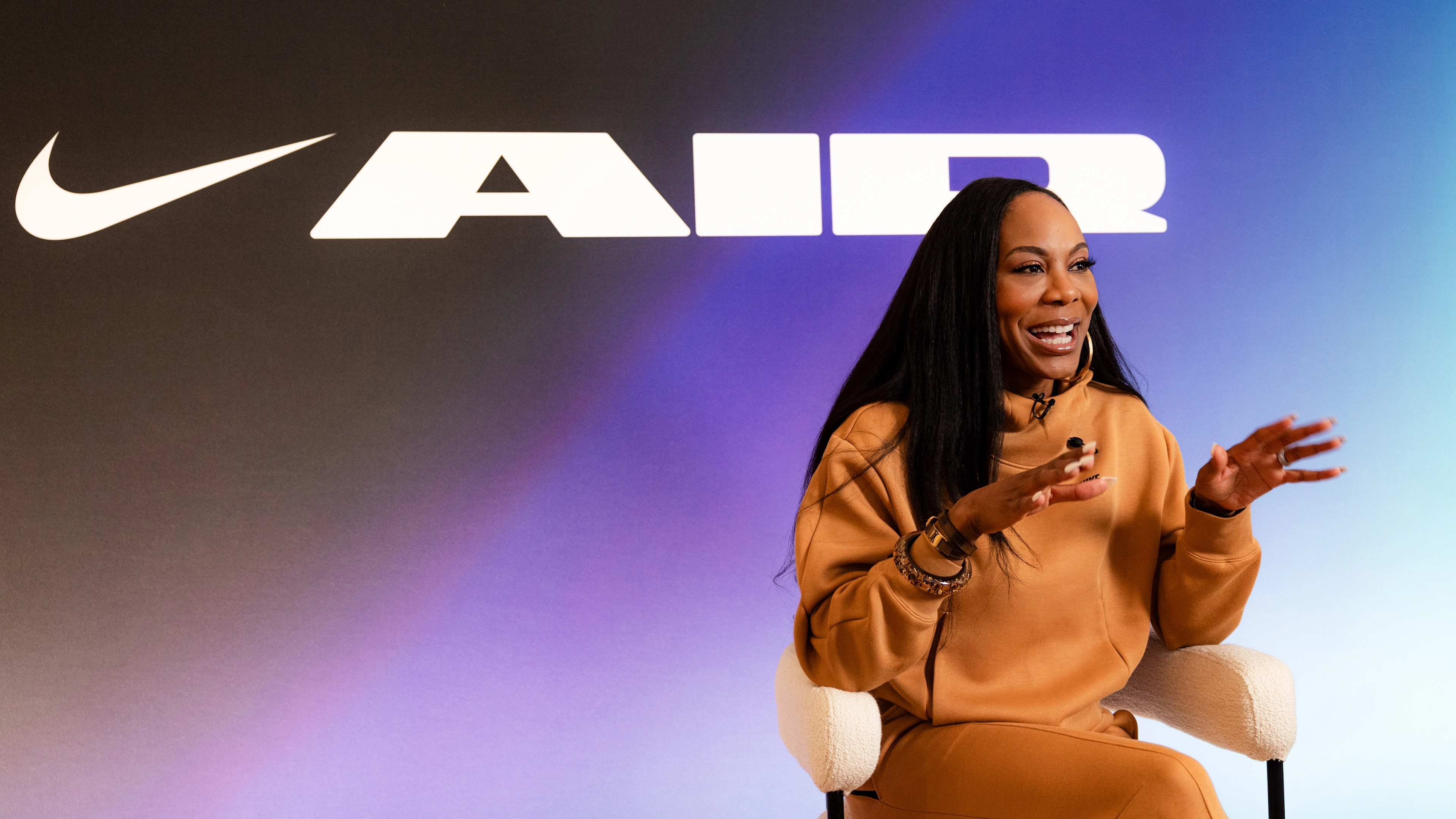 Front angled view of Sanya Richards-Ross wearing Nike Sportswear Tech Fleece in the Flax color. Sanya sits on a white stool, against a blue & purple gradient background with a large white logo for Nike AIR.