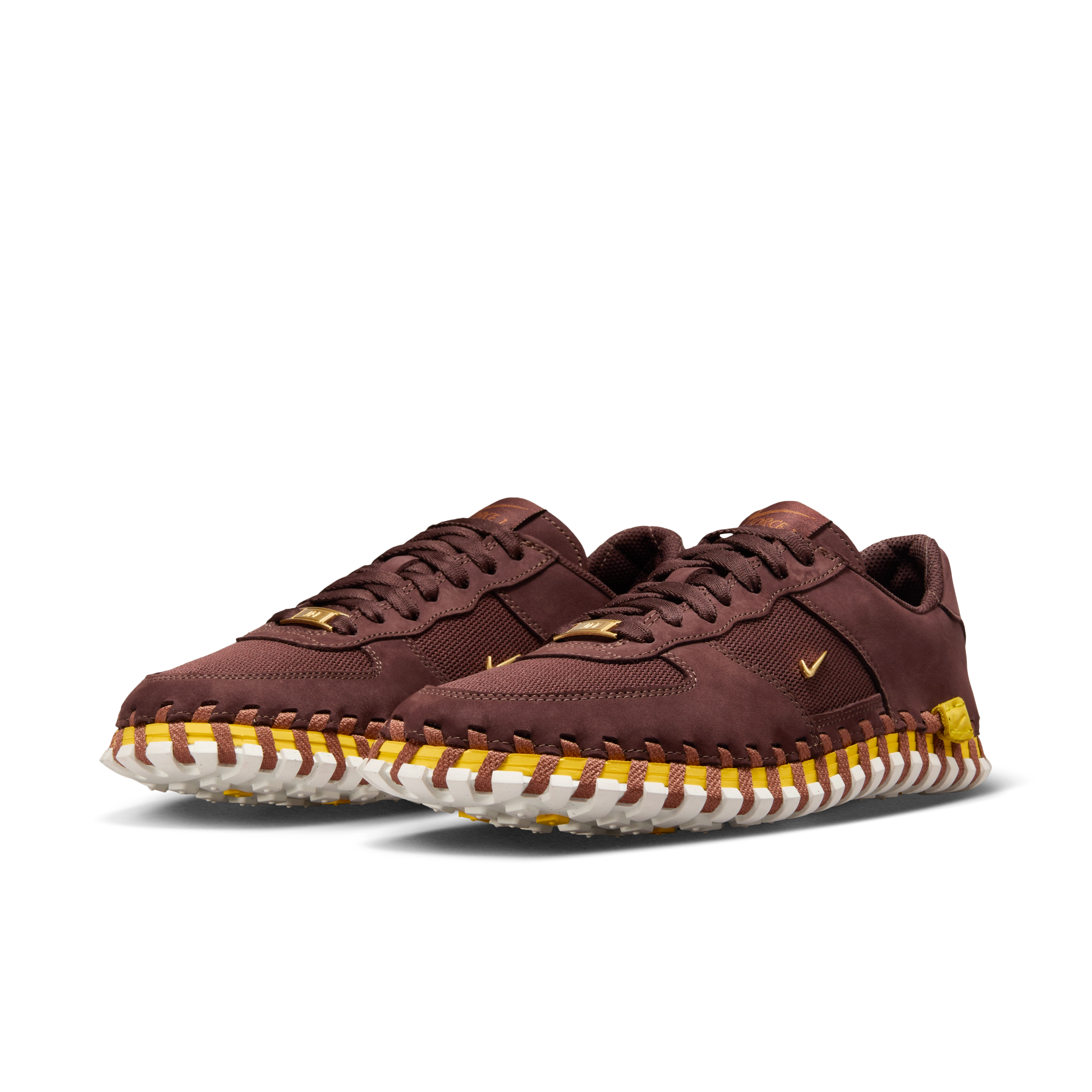 Front angled view of a pair of Nike x Jacquemus J Force 1 Low LX SP with Earth brown upper, tour yellow midsole, and white sole. Includes, brown laces and AF1 lace tags in gold metal. There are small metallic gold Swooshes on the sides of the shoes. The pair floats on a black background.