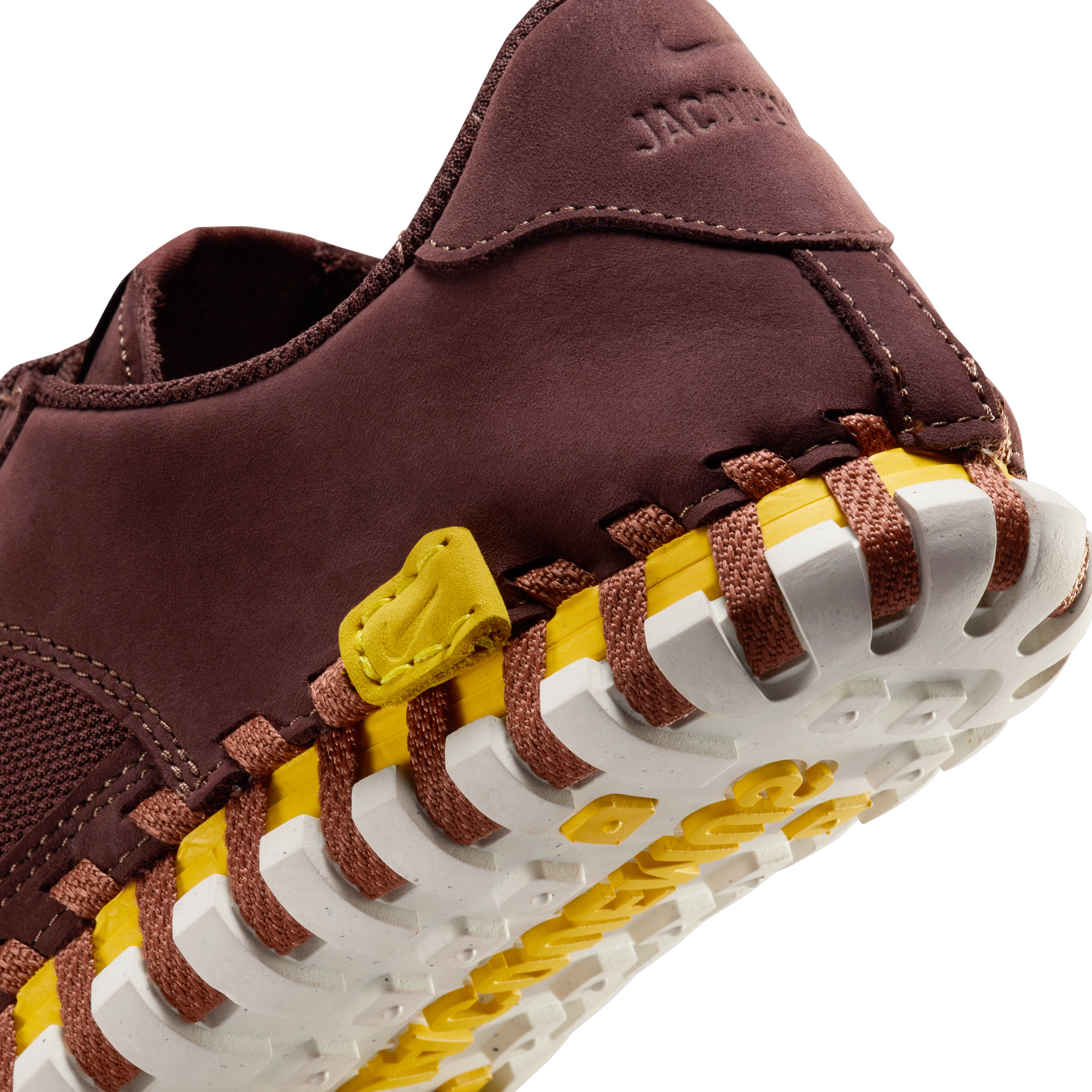 Back detail view of a Nike x Jacquemus J Force 1 Low LX SP with Earth brown upper, tour yellow midsole, and white sole. There is a small sewn-in yellow leather label at the base of the heel with an embossed Swoosh, and an embossed Jacquemus logo on the heel cup. The shoe floats on a black background.