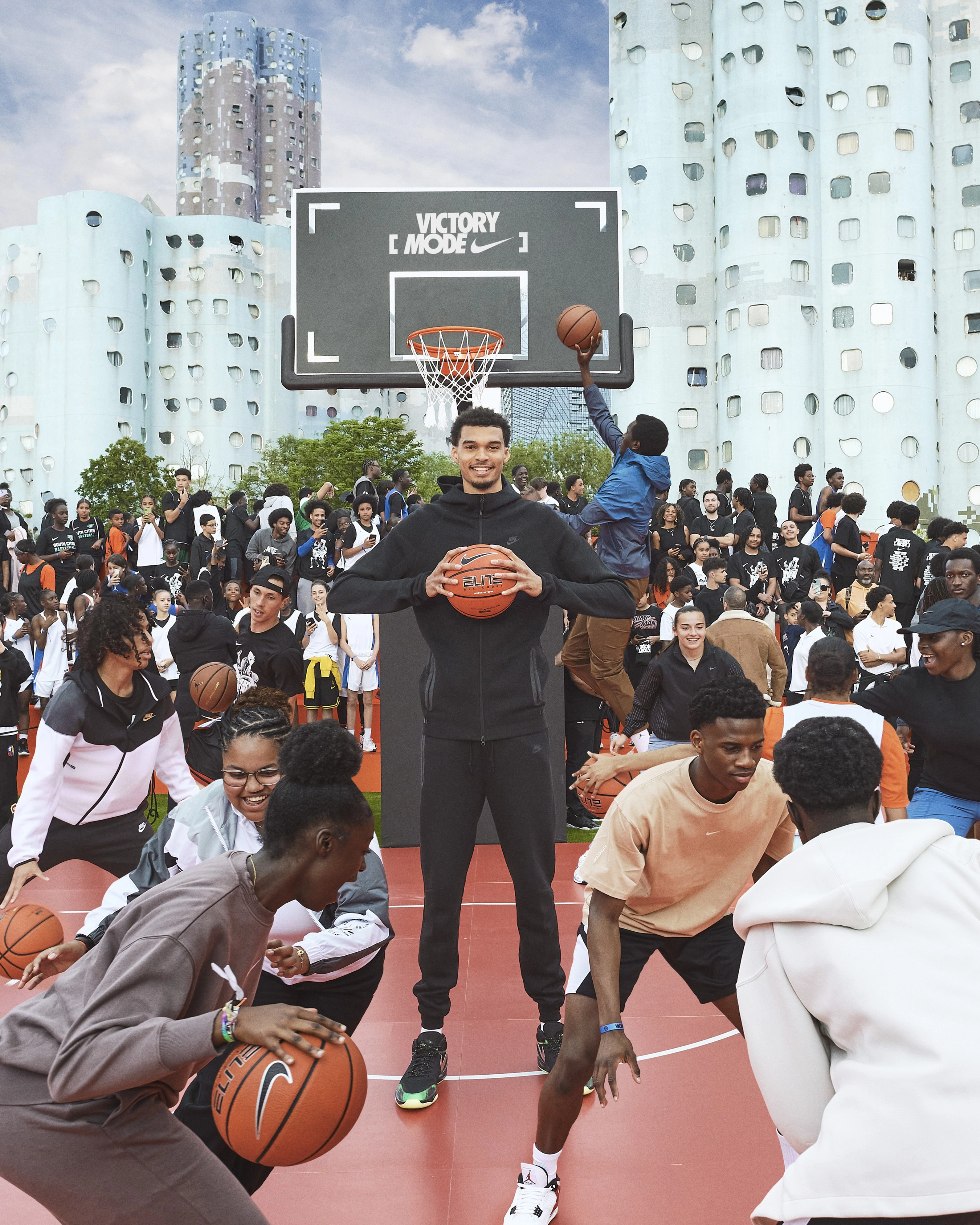 Front view of Victor Wembanyama, wearing black Nike Tech Fleece, smiling as he holds a basketball surrounded by a crowd of kids playing basketball at the Victory Mode event in Nanterre, France.