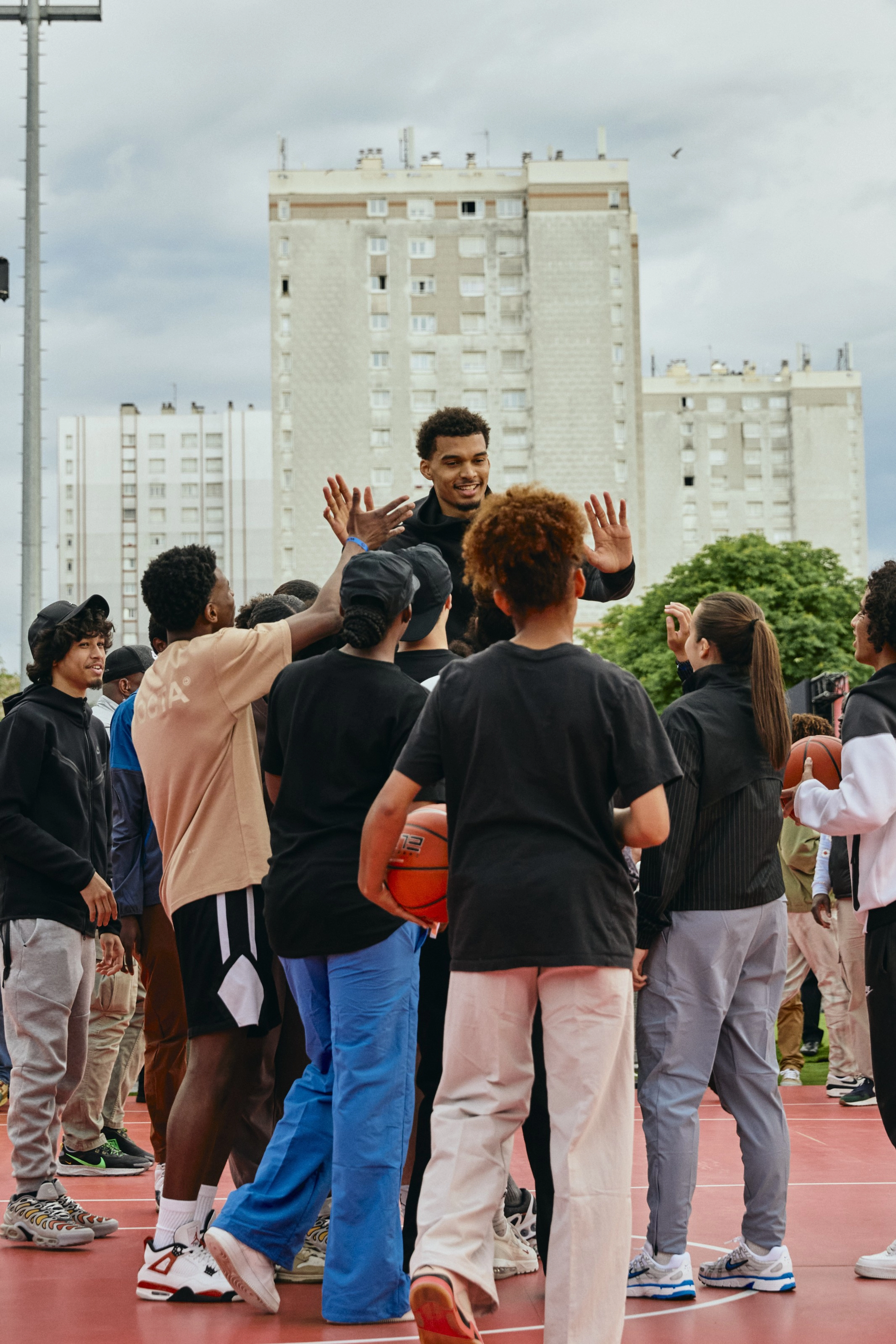 Victor Wembanyama gives high fives to a crowd of young athletes at the Victory Mode event in Nanterre, France.  They stand on a red court with white buildings in the background.