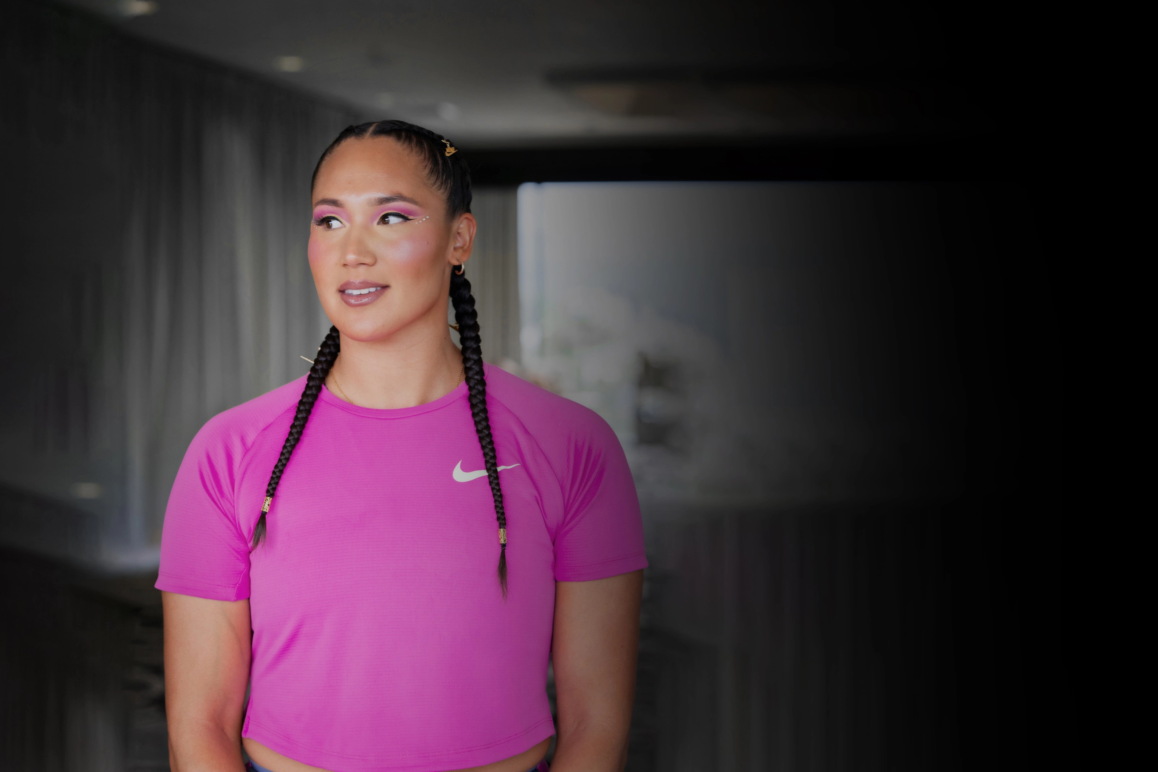 Portrait of American track and field athlete, Janne Kassanavoid, wearing a pink Nike Dri-FIT tee with matching eyeshadow. She stands against a blurred dark background.