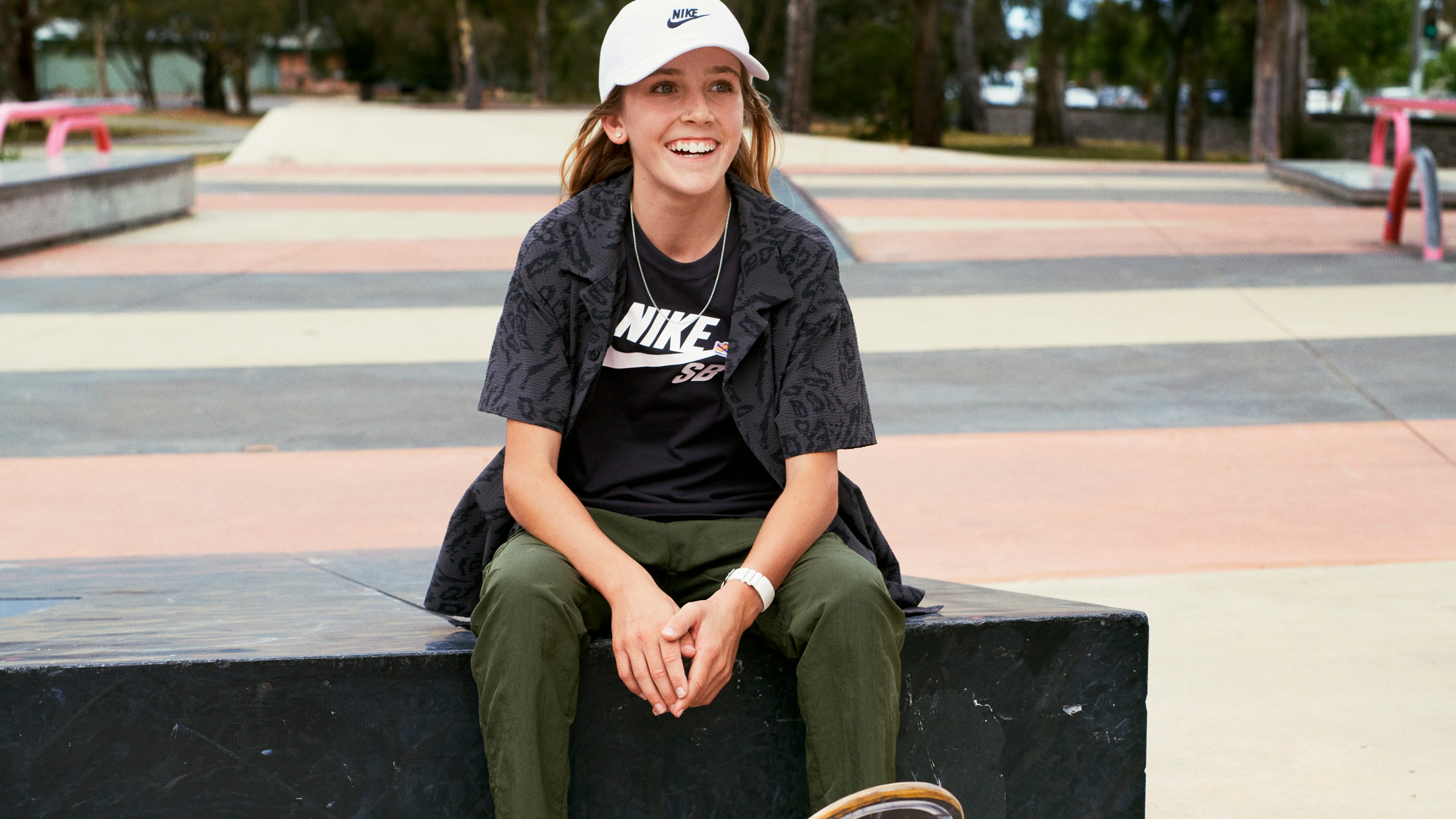 Chloe Covell smiling and wearing a white Nike baseball cap and black Nike t-shirt sitting on a concrete wall at a skate park.