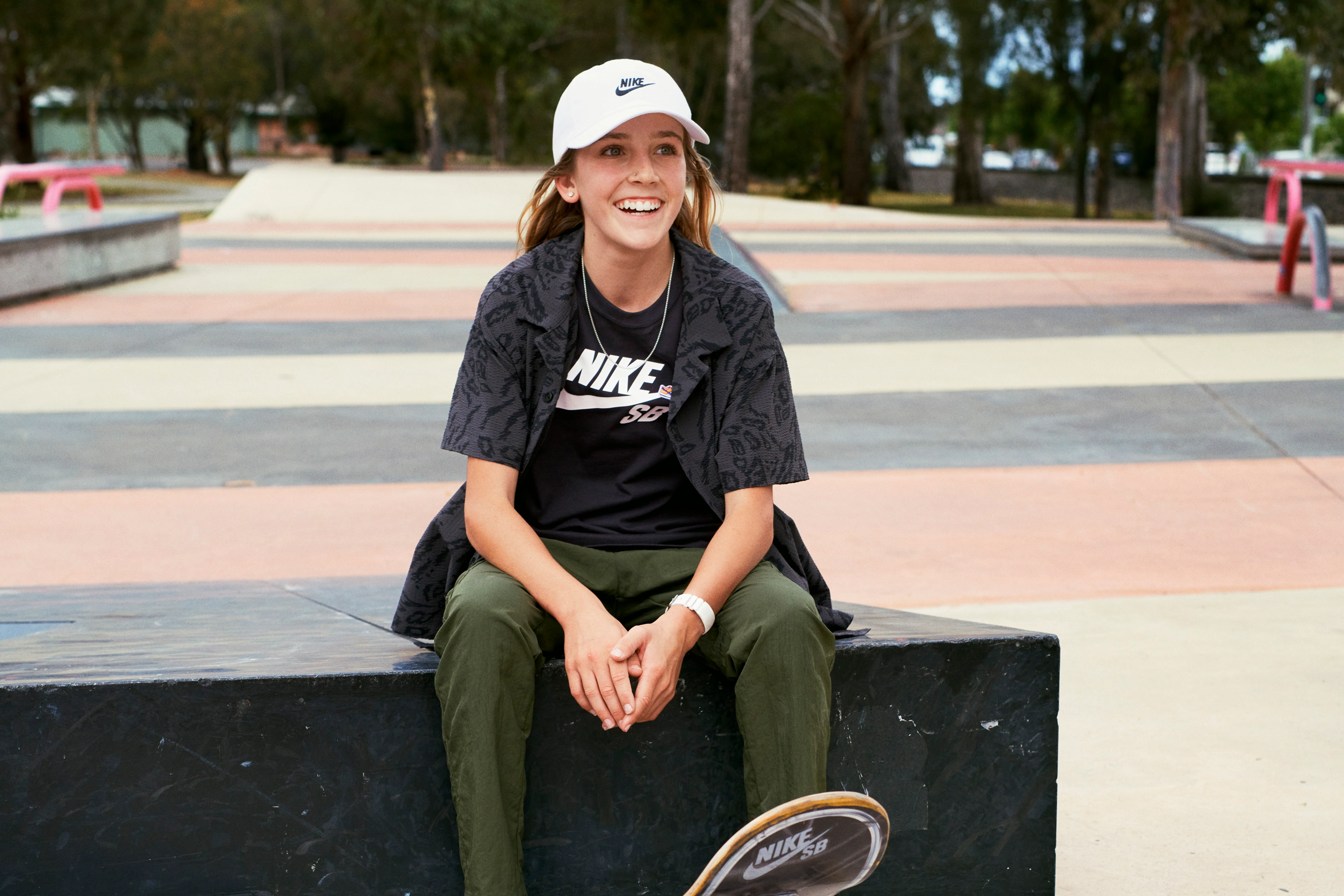 Chloe Covell smiling and wearing a white Nike baseball cap and black Nike t-shirt sitting on a concrete wall at a skate park.