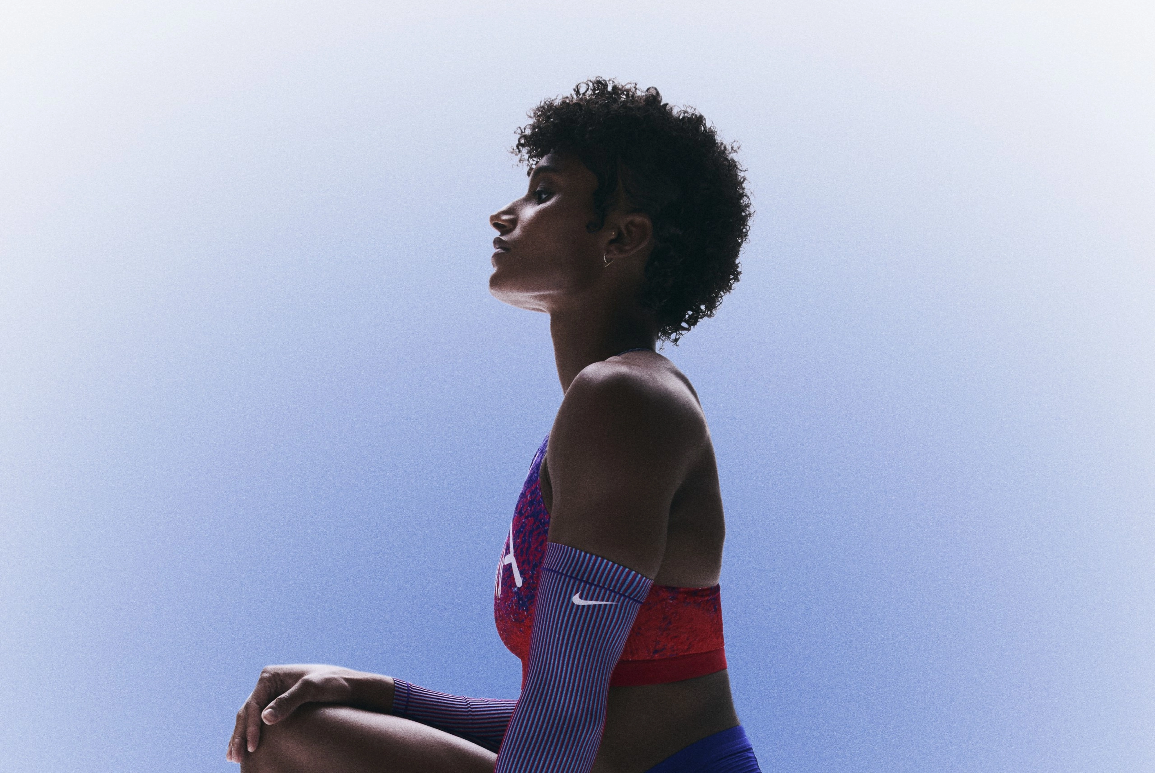 Side view of Anna Cockrell, kneeling on one knee in her Team USA uniform. She holds her head high, facing left against a blue gradient background.