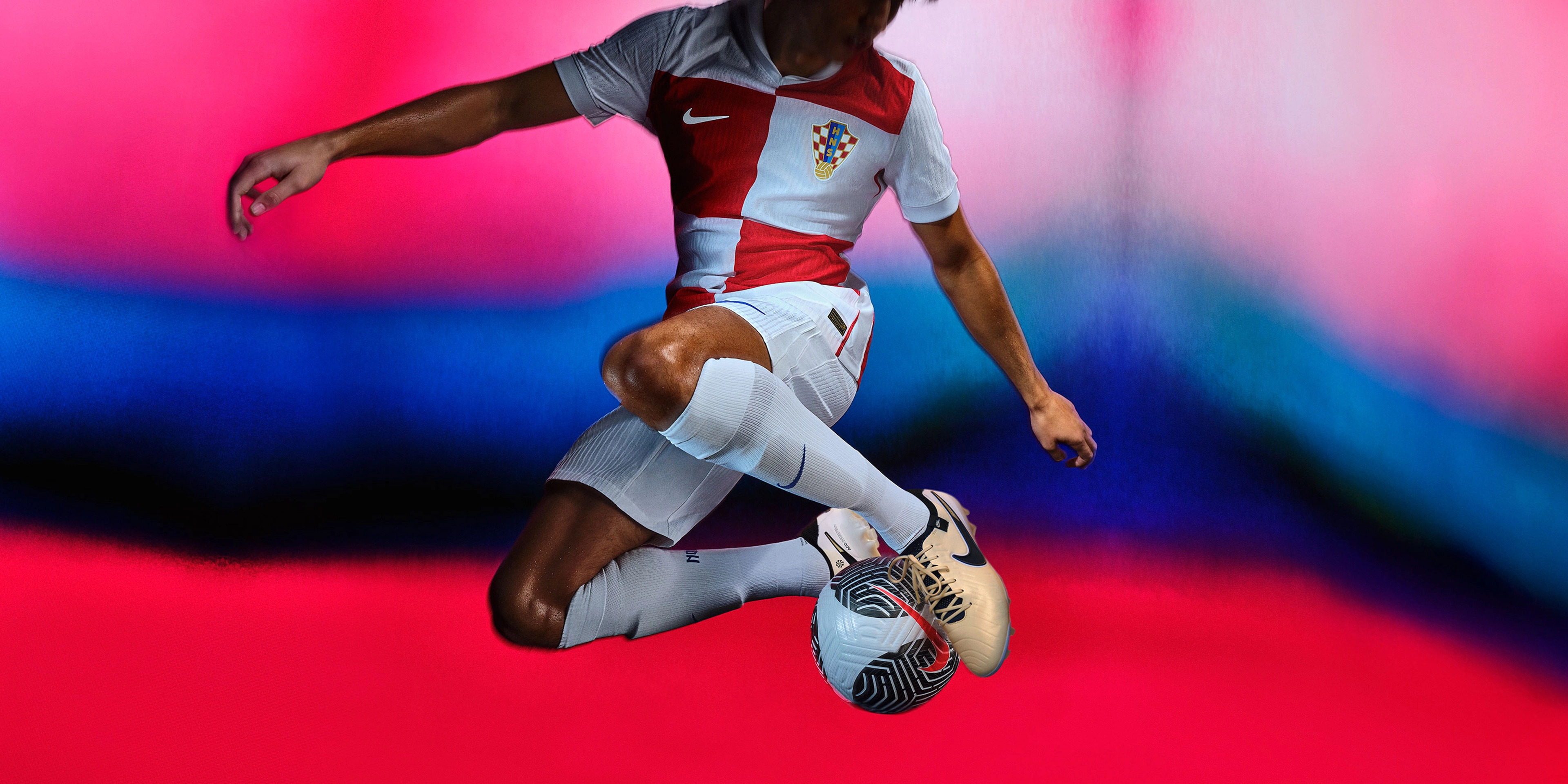 Athlete in a jumping pose with a soccer ball between their feet wearing a soccer jersey with a large red and white square pattern, as well as matching white shorts. They are also wearing white socks.