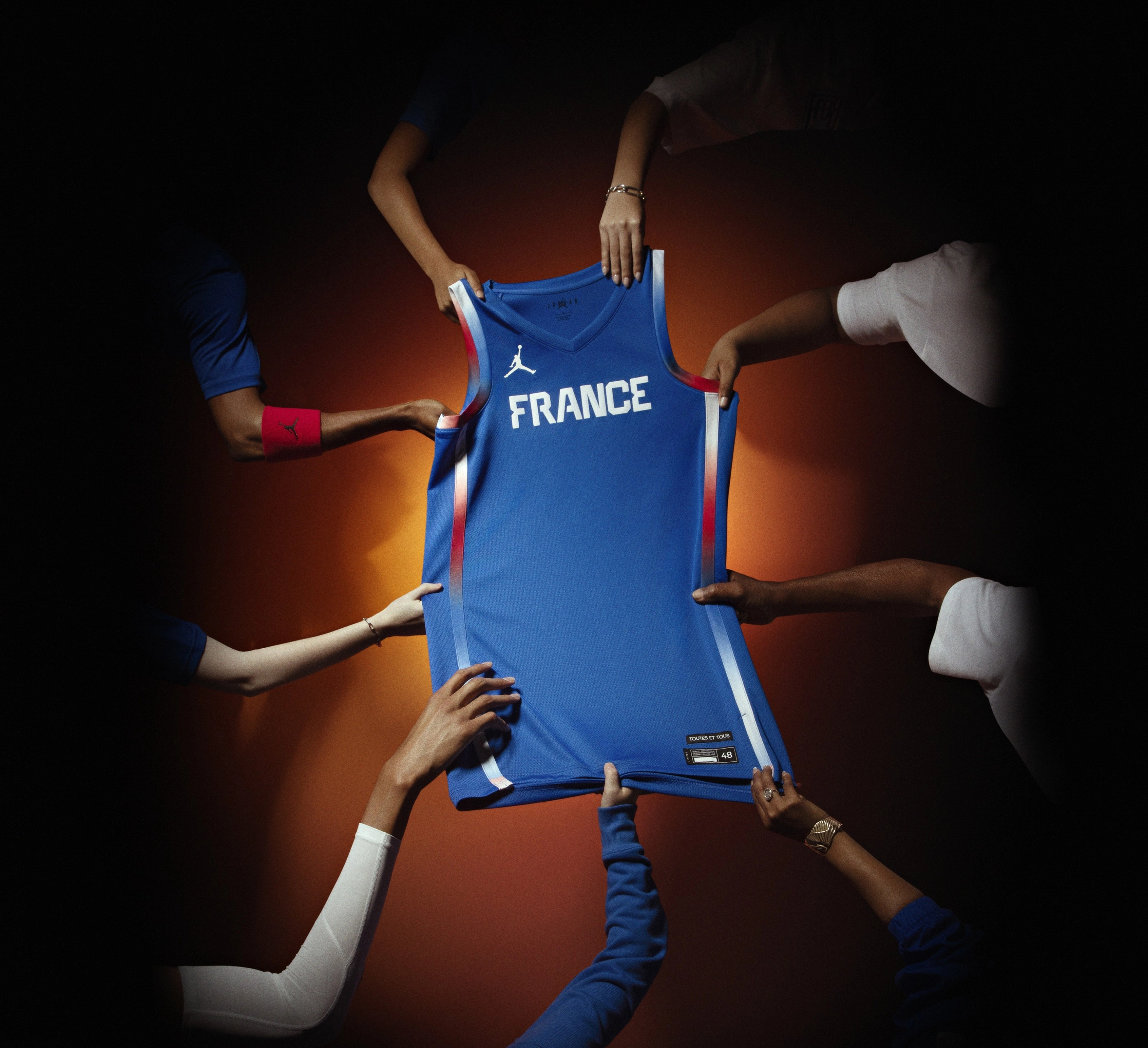 Several hands collectively hold up the sky blue France Basketball National Team Jersey, which has the word "France" prominently featured on the front.