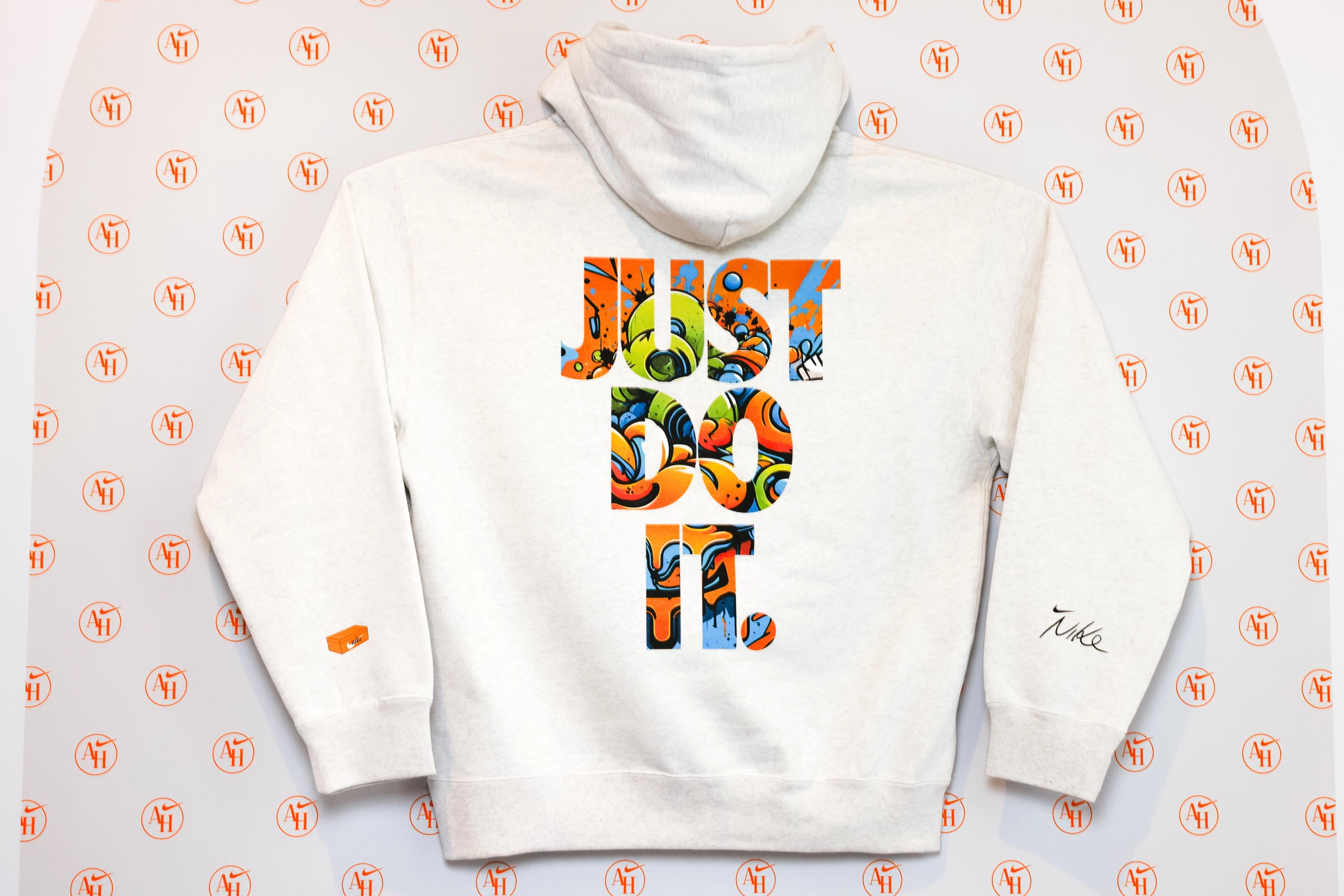Back view of a white hoodie that has been customized in the AI space in the Nike Athlete House. A large, colorful JUST DO IT screenprint appears on the back of the sweatshirt, while the sleeves display a small orange footwear box and a scripted Nike logo. The hoodie sits on top of a repeating AH logo background.
