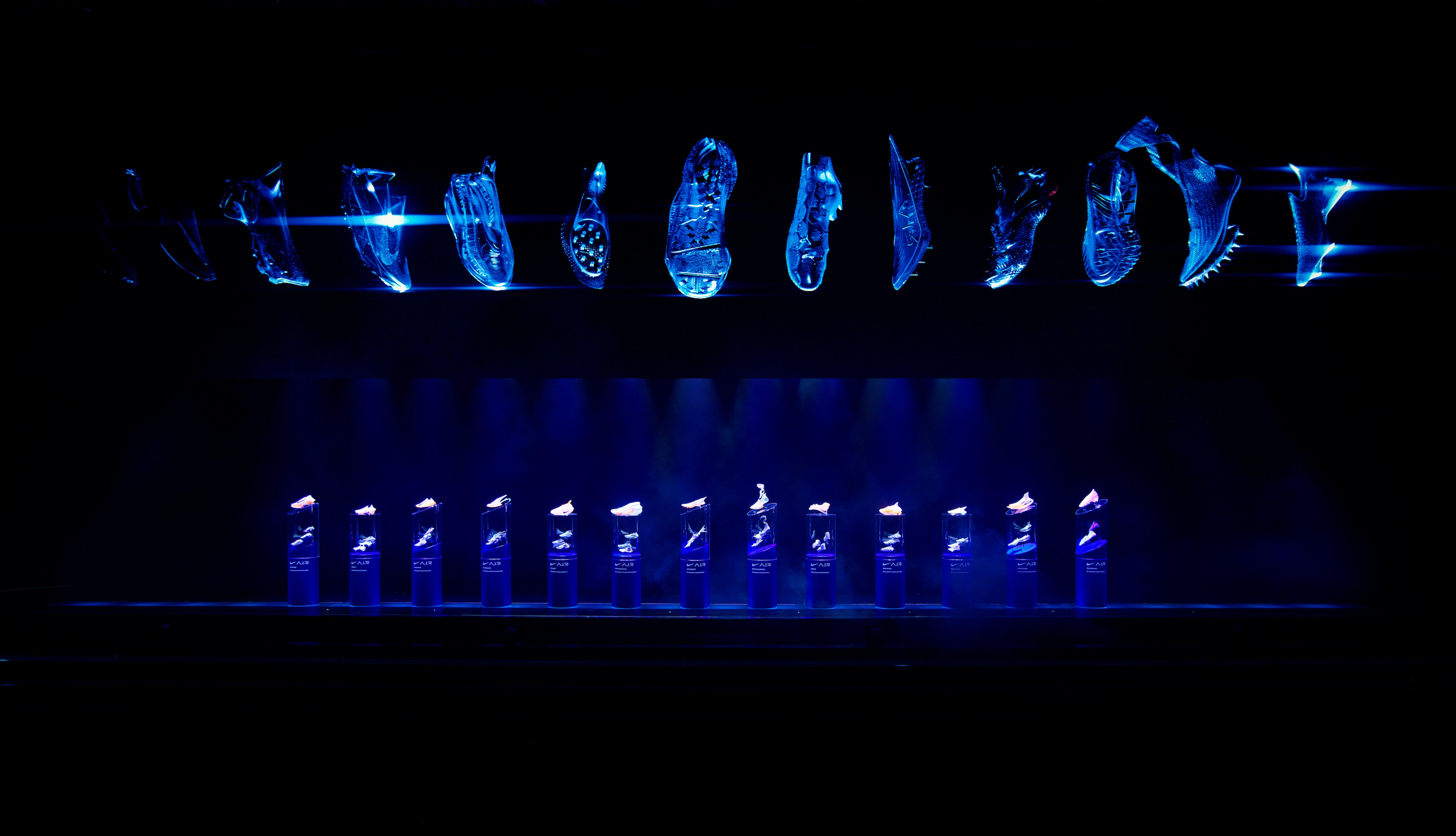 A stage featuring a row of pedestals displaying Nike sneakers in navy blue lighting. The sneakers are displayed in bright blue on the overhead screen.