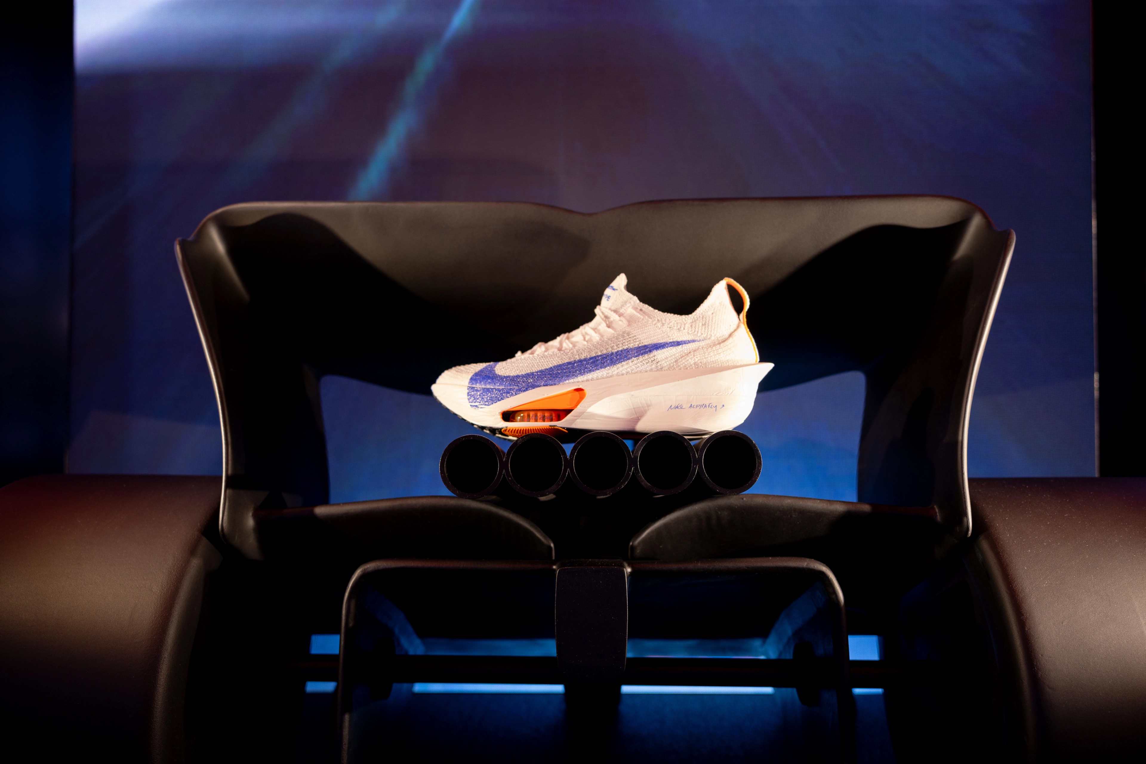 A white Nike sneaker with a navy blue swoosh on the side and a neon orange sole support on top of a display.