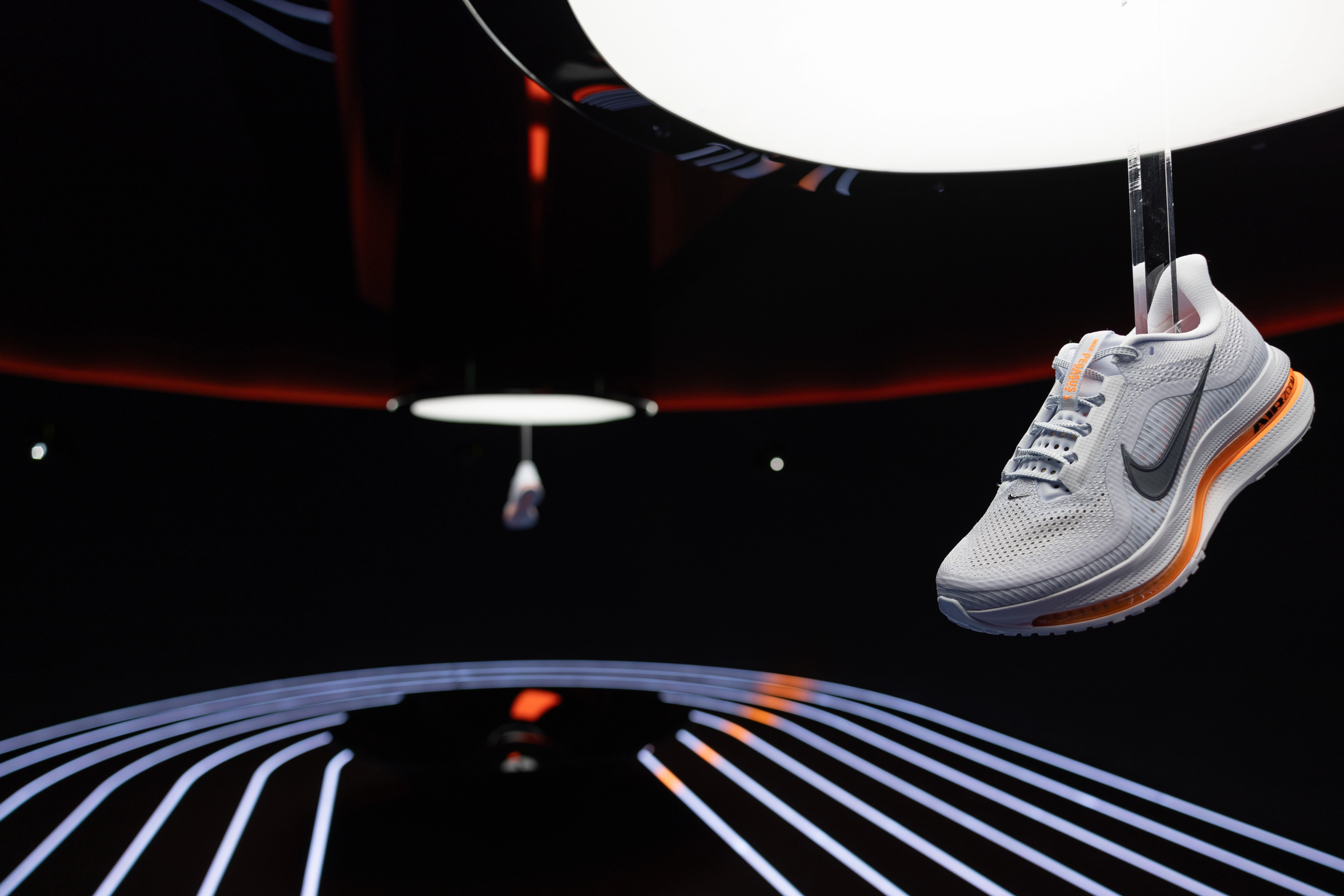 A close up view of a Nike shoe hanging from a low ceiling in a dark room. Another slightly blurred sneaker can be seen in a similar display behind it.