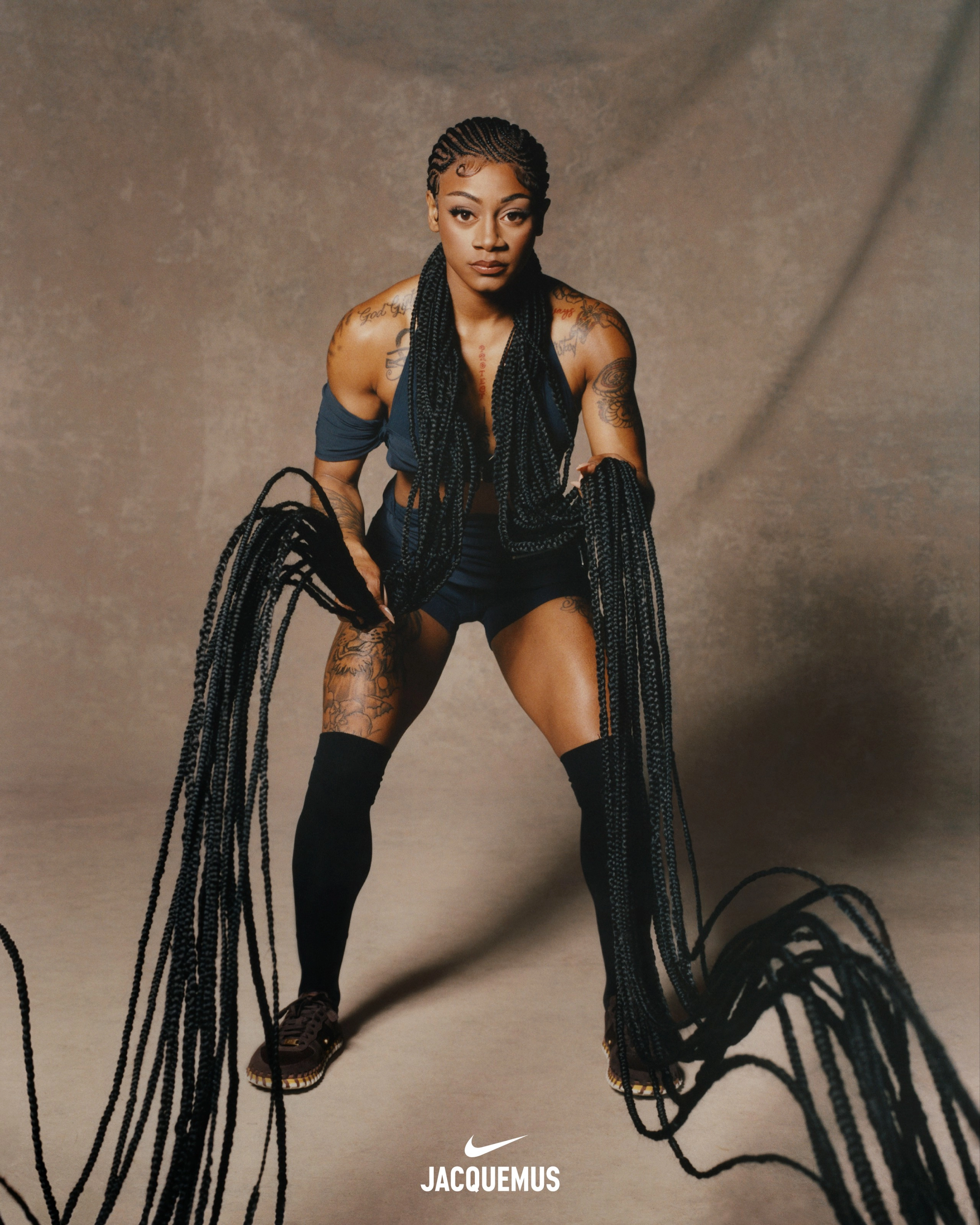 Front, full-body view of Sha'Carri Richardson wearing Nike X Jacquemus' layered shorts and halter neck sleeveless crop top in dark obsidian, over-the-knee socks and the Nike J Force 1 Low LX SP shoes in brown. Sha'Carri's hair is in ornate cornrows and she uses her extended braids like she's exercising with heavy ropes. You can see tattoos on her chest, arms, and legs. Sha'Carri looks directly at the camera, not smiling, as she stands in front of a beige material background.