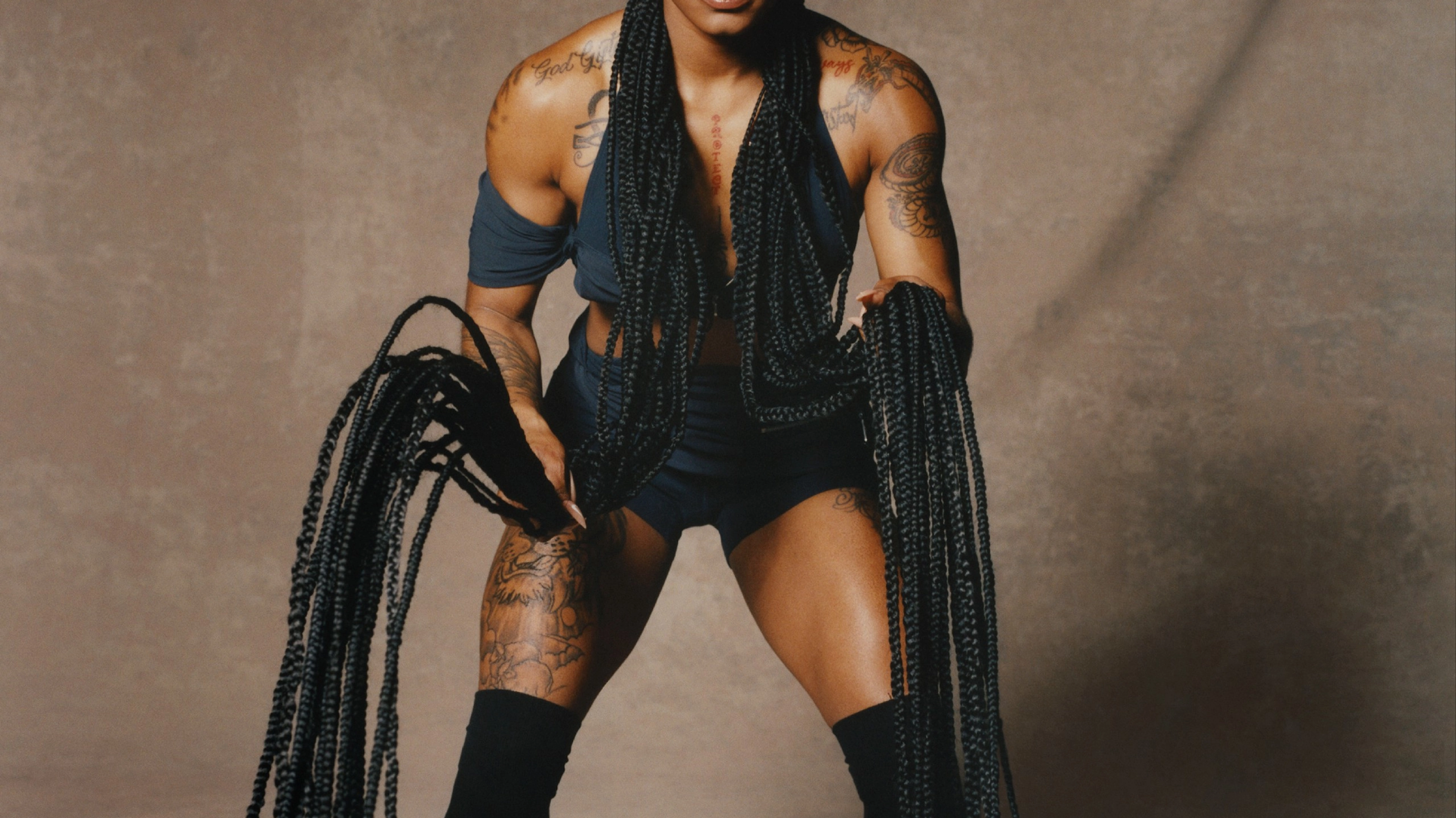 Front, full-body view of Sha'Carri Richardson wearing Nike X Jacquemus' layered shorts and halter neck sleeveless crop top in dark obsidian, over-the-knee socks and the Nike J Force 1 Low LX SP shoes in brown. Sha'Carri's hair is in ornate cornrows and she uses her extended braids like she's exercising with heavy ropes. You can see tattoos on her chest, arms, and legs. Sha'Carri looks directly at the camera, not smiling, as she stands in front of a beige material background.
