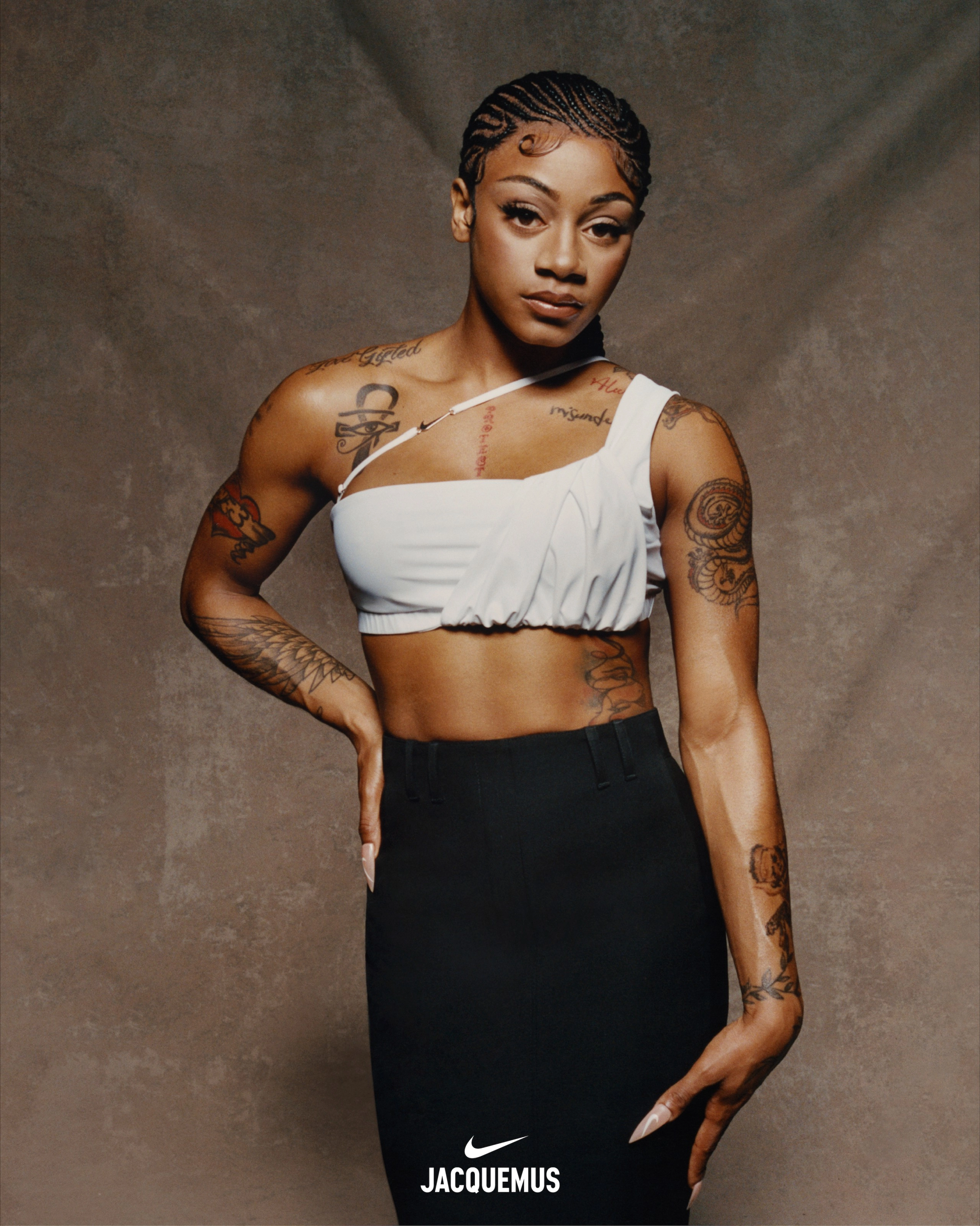 Close up of Sha'Carri Richardson wearing Nike X Jacquemus' slightly ruched white cropped one-shoulder tank and a high-waist maxi-length black skirt. Sha'Carri's hair is in ornate cornrows and we see tattoos on her chest and arms. Sha'Carri looks directly at the camera, not smiling, holding one hand at her hip and the other  at her thigh.  She stands in front of a beige material background.