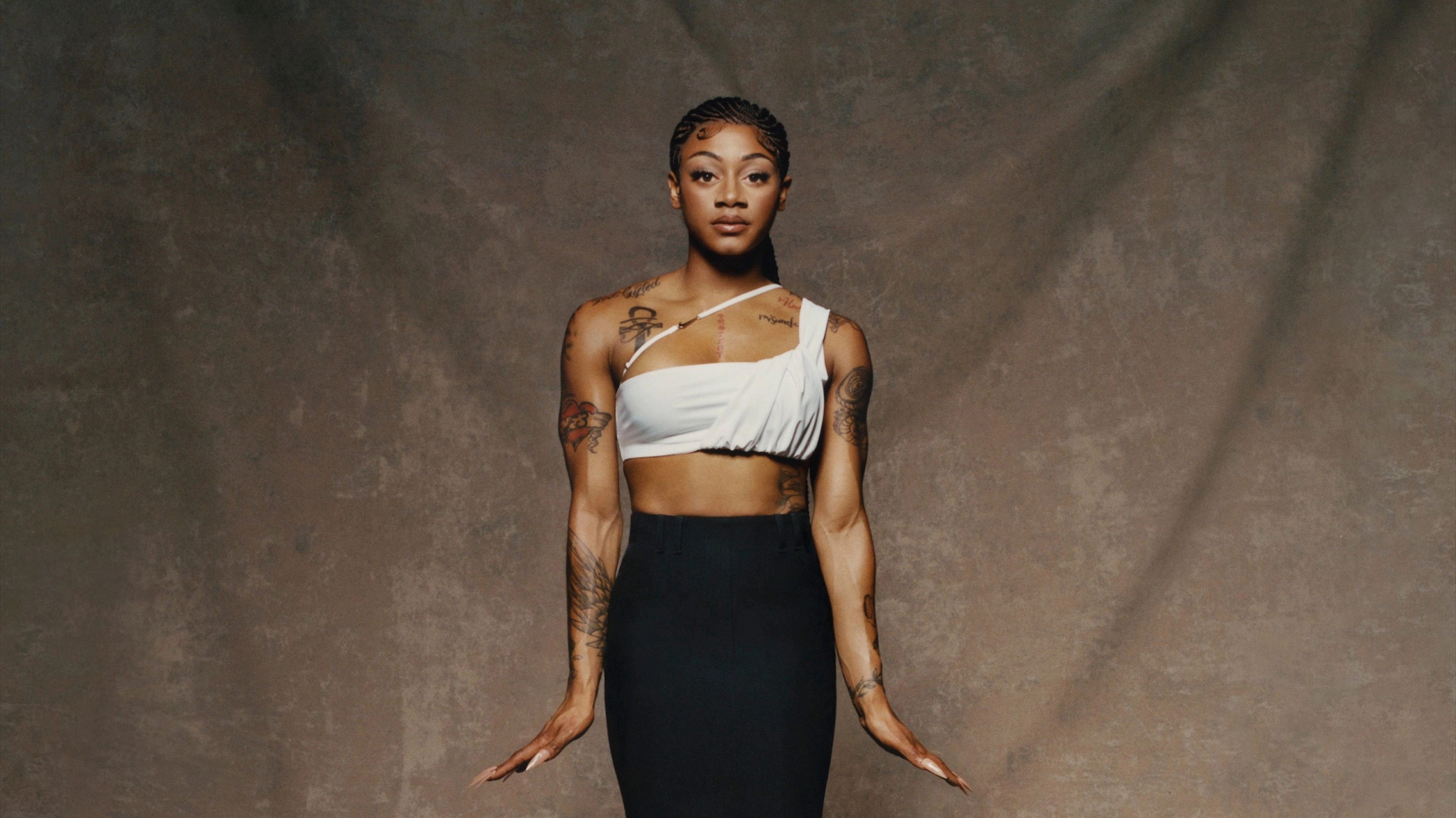 Front, full-body view of Sha'Carri Richardson wearing Nike X Jacquemus' slightly ruched white cropped one-shoulder tank and a high-waist maxi-length black skirt. Sha'Carri's hair is in ornate cornrows and extended braids pool around her on the floor. You can see tattoos on her chest and arms. Sha'Carri looks directly at the camera, not smiling, holding her hands outwards at her thighs, in front of a beige material background.