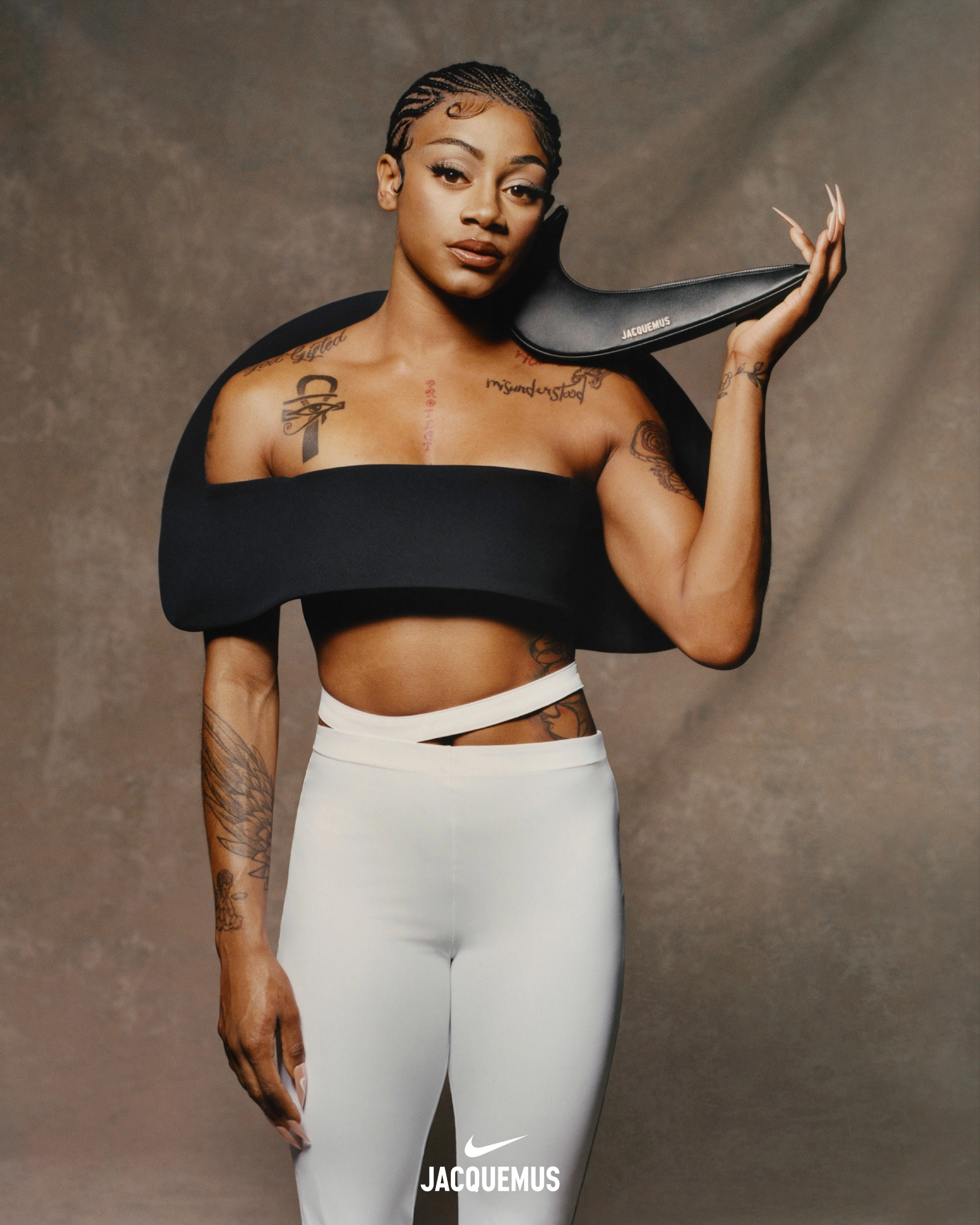 Front view of Sha'Carri Richardson wearing Nike x Jacquemus Women's Pants in white. She wears a sculptural black crop top, while holding one hand at her thigh while the other supports a black Nike x Jacquemus Le Swoosh bag on her shoulder. Sha'Carri's hair is in ornate cornrows. She looks directly at the camera as she stands in front of a beige material background.
