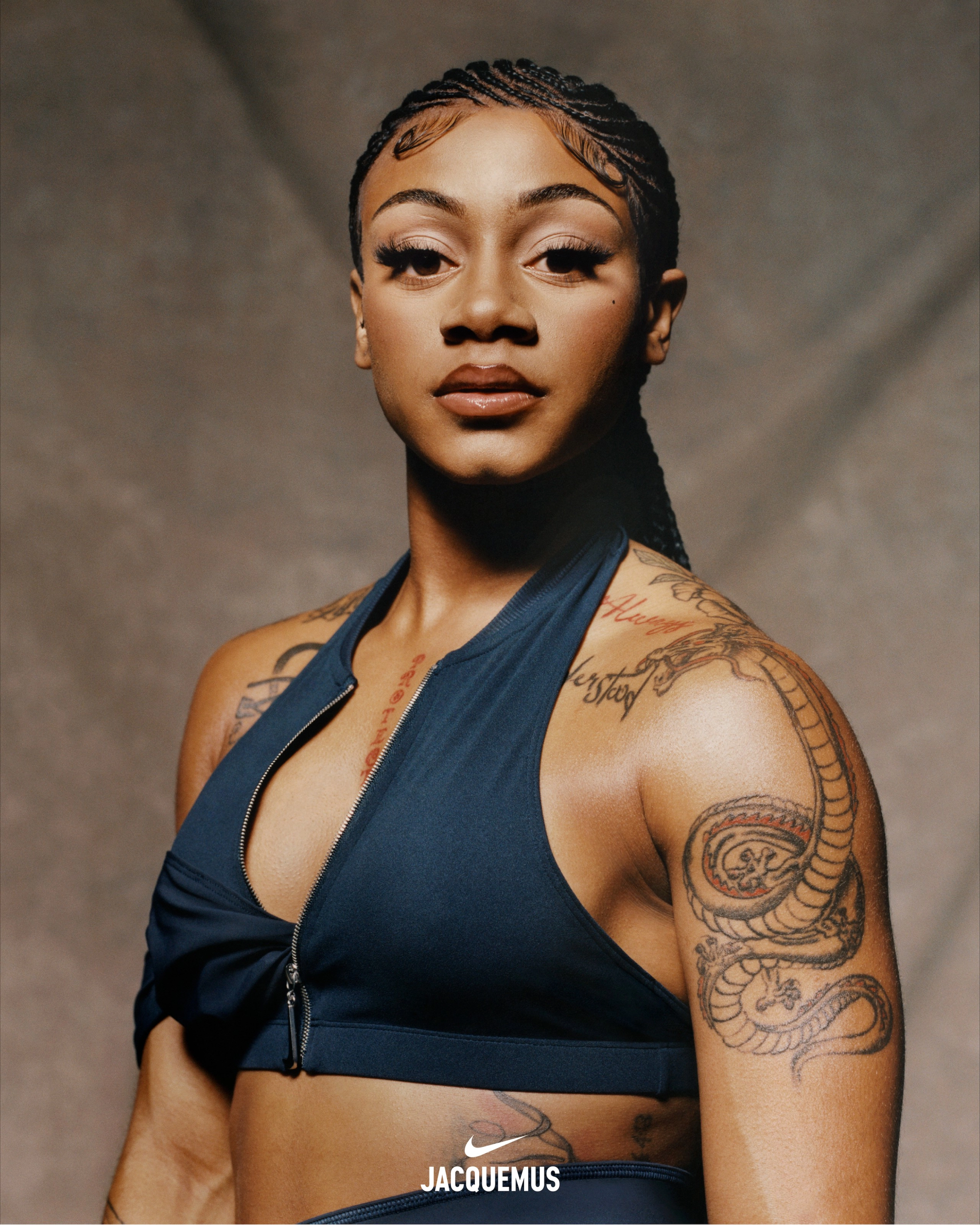Close up of Sha'Carri Richarson wearing a dark obsidian Nike x Jacquemus Halter Neck Sleeveless Crop Top. We see tattoos on her chest, arms and shoulders. Sha'Carri's hair is in ornate cornrows and she wears extended eyelashes. She looks directly at the camera against a beige material background.