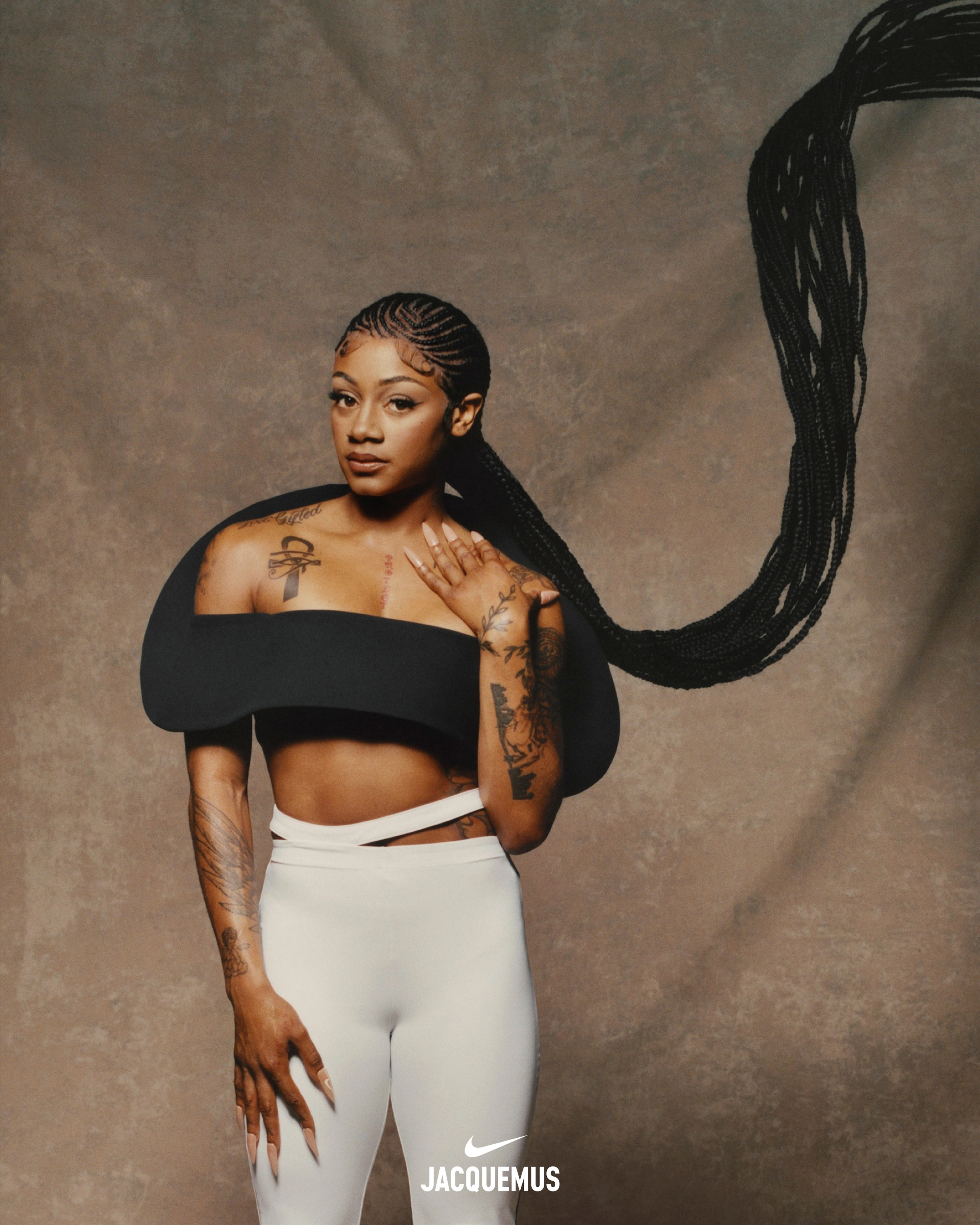 Front view of Sha'Carri Richardson wearing Nike x Jacquemus Women's Pants in white. She wears a sculptural black crop top, while holding one hand at her thigh and another at her shoulder. Sha'Carri's hair is in ornate cornrows and her extended braids fly upward to the right of her body. Sha'Carri looks directly at the camera as she stands in front of a beige material background.